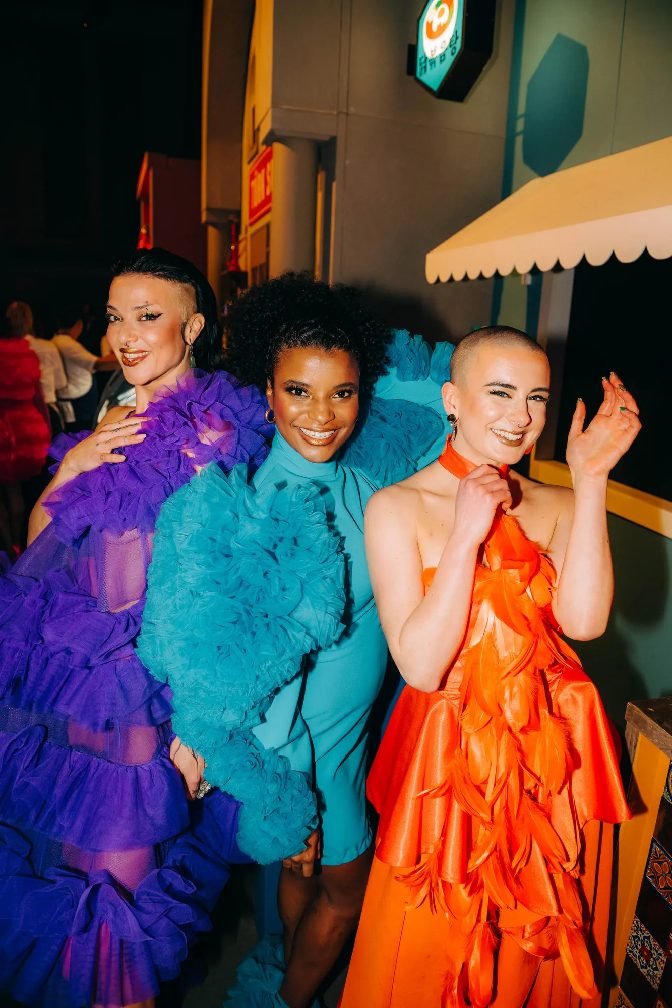 Three women smiling and posing in colorful ruffled dresses, one in purple, one in blue, and one in orange featuring feather details.