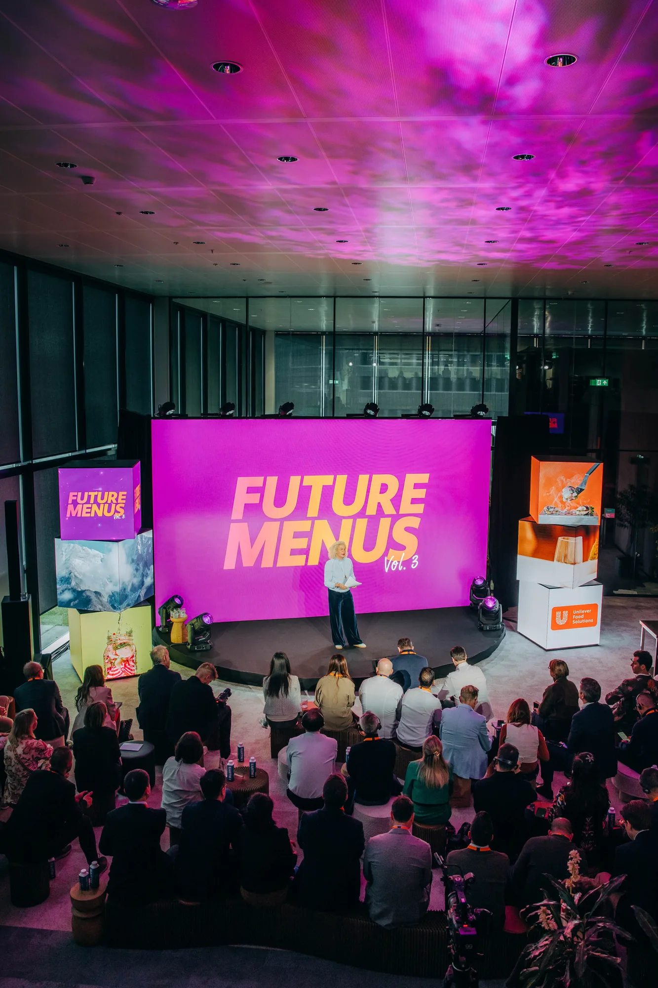 Speaker on stage presenting at an event with a large screen behind displaying 'FUTURE MENUS Vol. 3' in yellow text on a purple background, attended by a seated audience.