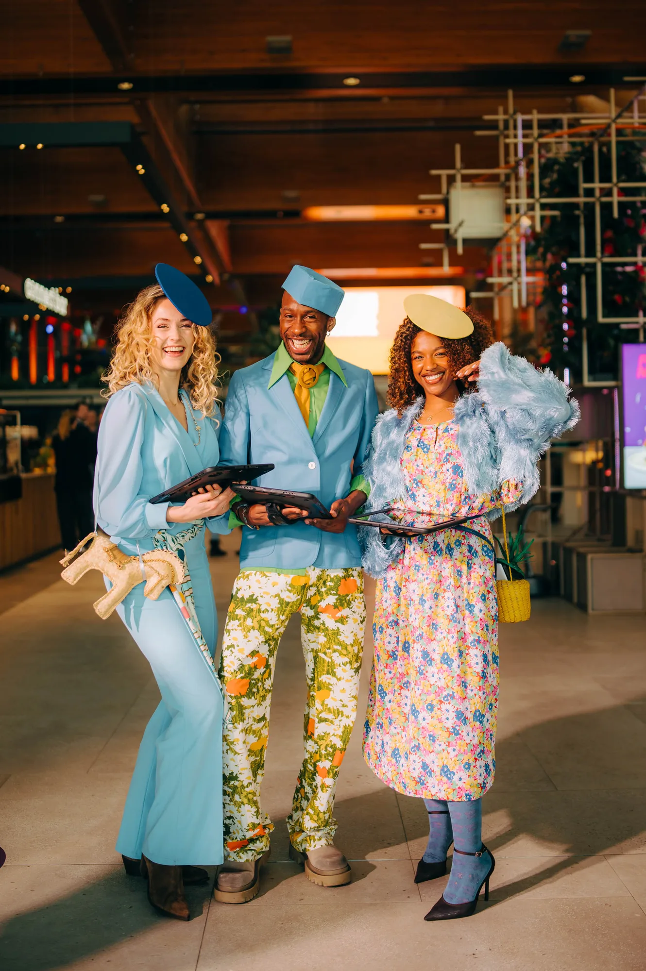 Three stylishly dressed people smiling indoors, wearing colorful and vintage-inspired outfits with small hats and holding tablets.