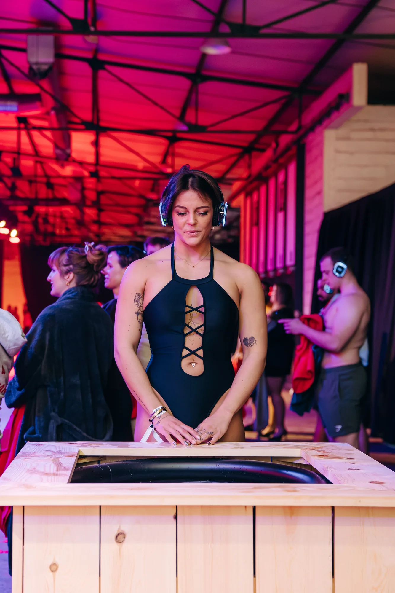 Woman in a black swimsuit wearing headphones, standing at a wooden table in a room with pink and orange lighting and people in the background.