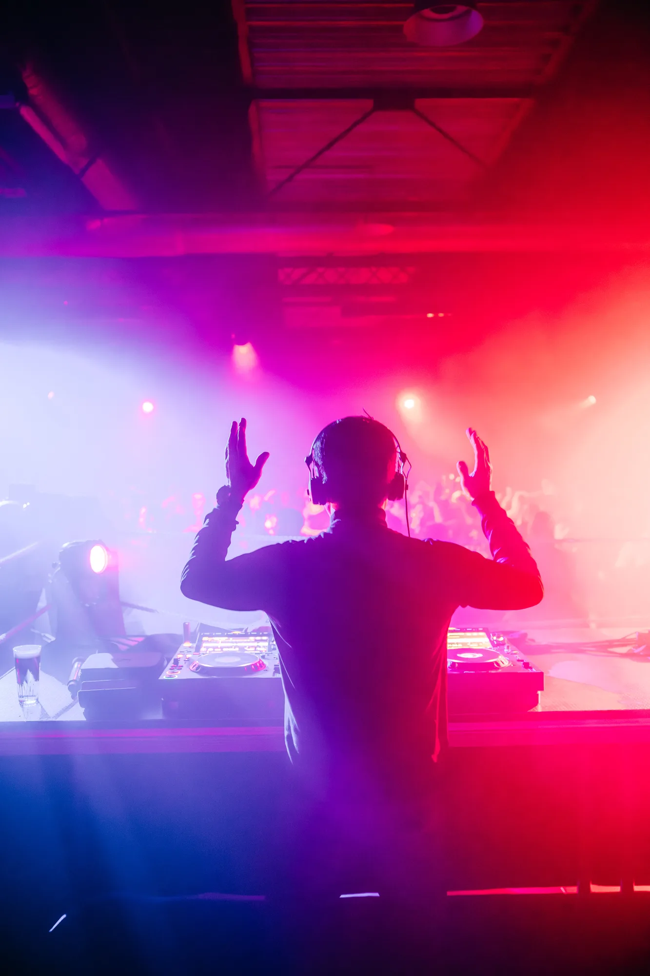 DJ wearing headphones with raised hands performing in front of a vibrant crowd under pink and purple stage lights.