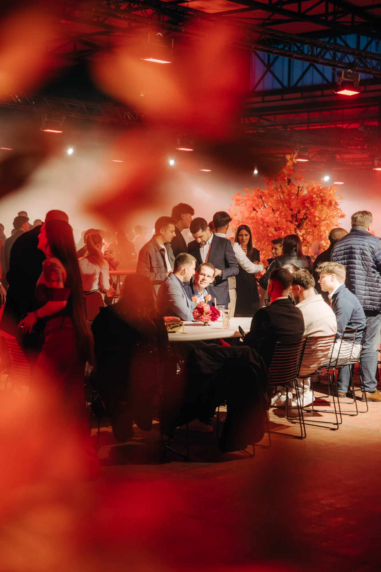 Group of people socializing and sitting at tables under warm orange lighting with decorative orange leaves in the background.