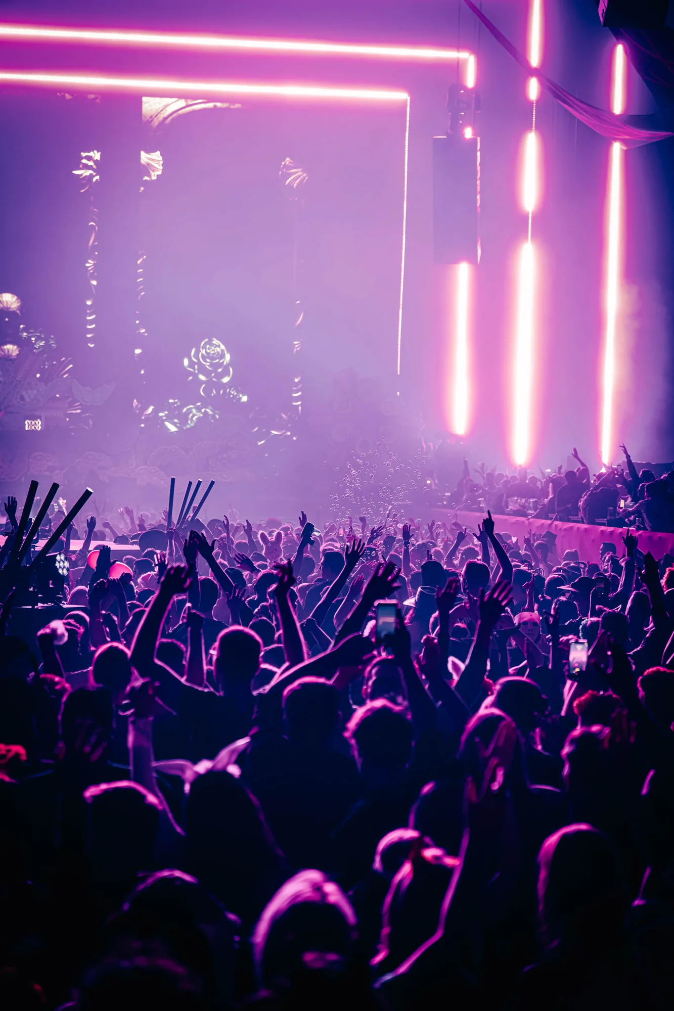 Crowd of people with raised hands enjoying a nightclub or concert under pink and purple neon lights.