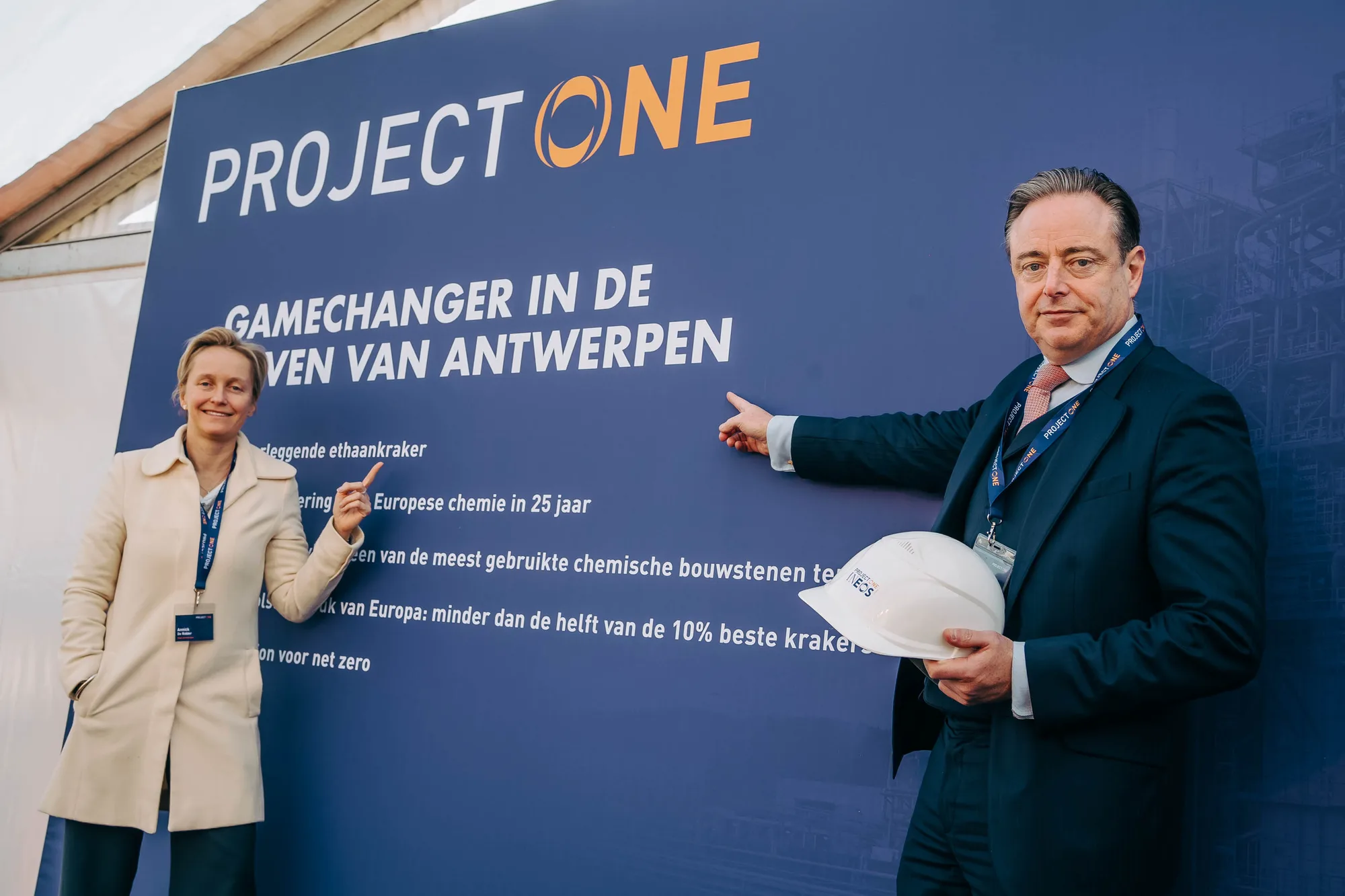Two professionally dressed people pointing at a blue signboard with the text 'PROJECT ONE' and Dutch information about a game-changing project in Antwerp.