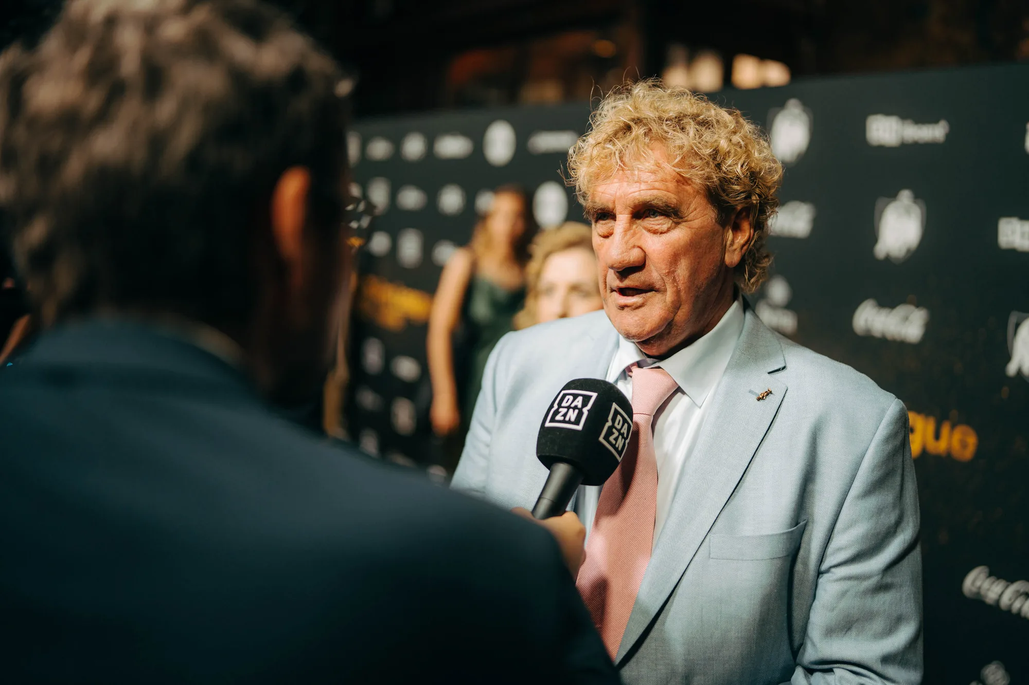 A man in a light blue suit and pink tie speaking into a DAZN microphone during an interview on a red carpet event.