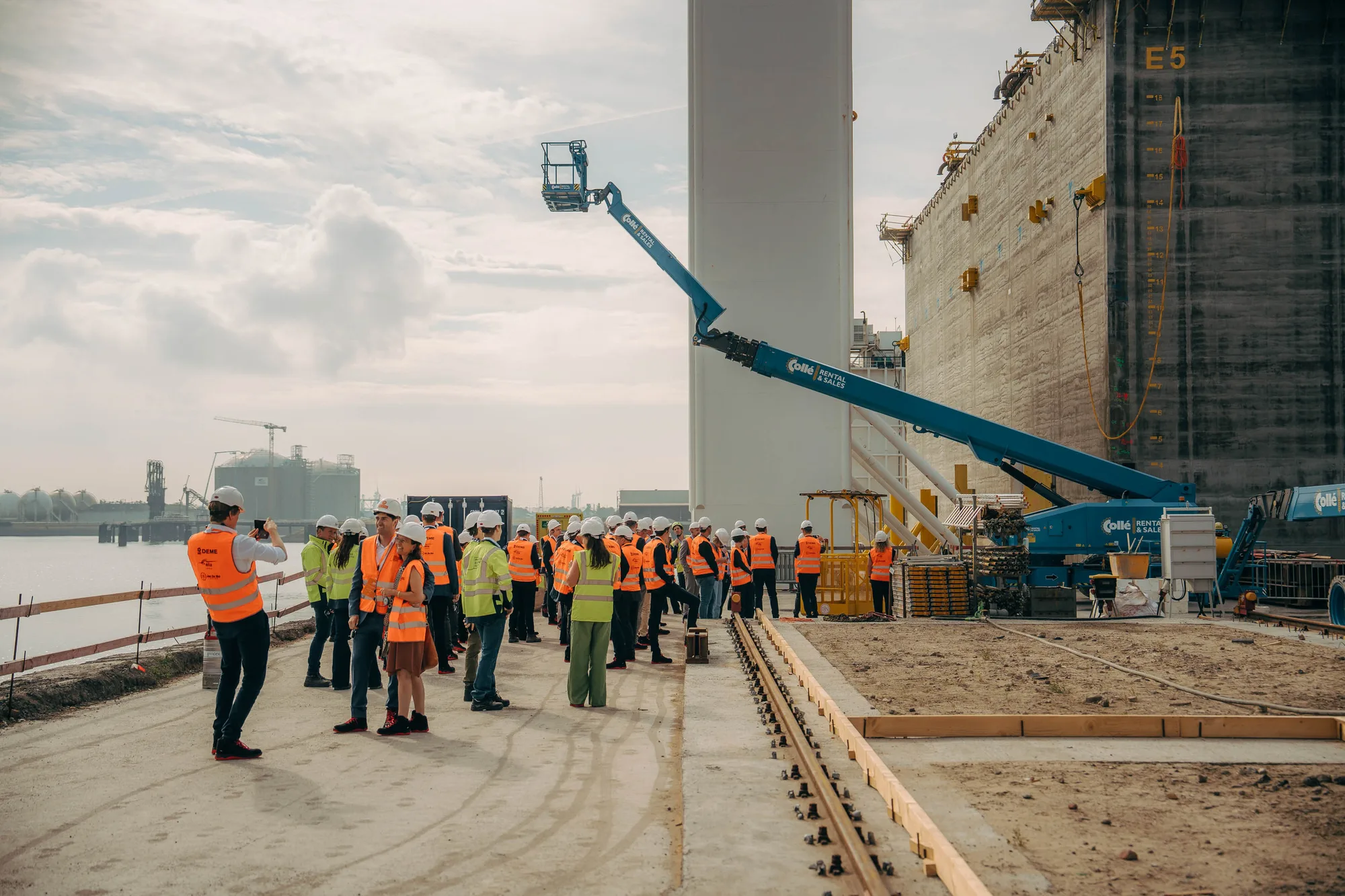 Group of construction workers wearing safety helmets and orange and yellow vests gathered at an industrial site near water with heavy machinery and large concrete structures.