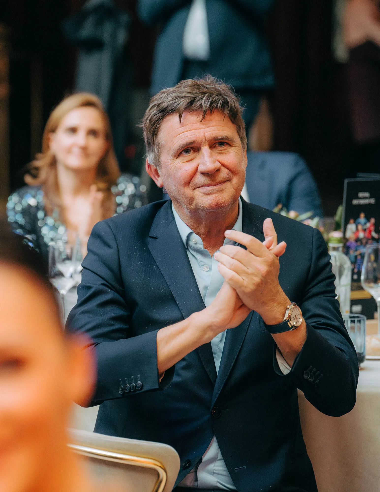 Middle-aged man in a dark suit clapping hands while seated at a formal event with blurred guests in the background.
