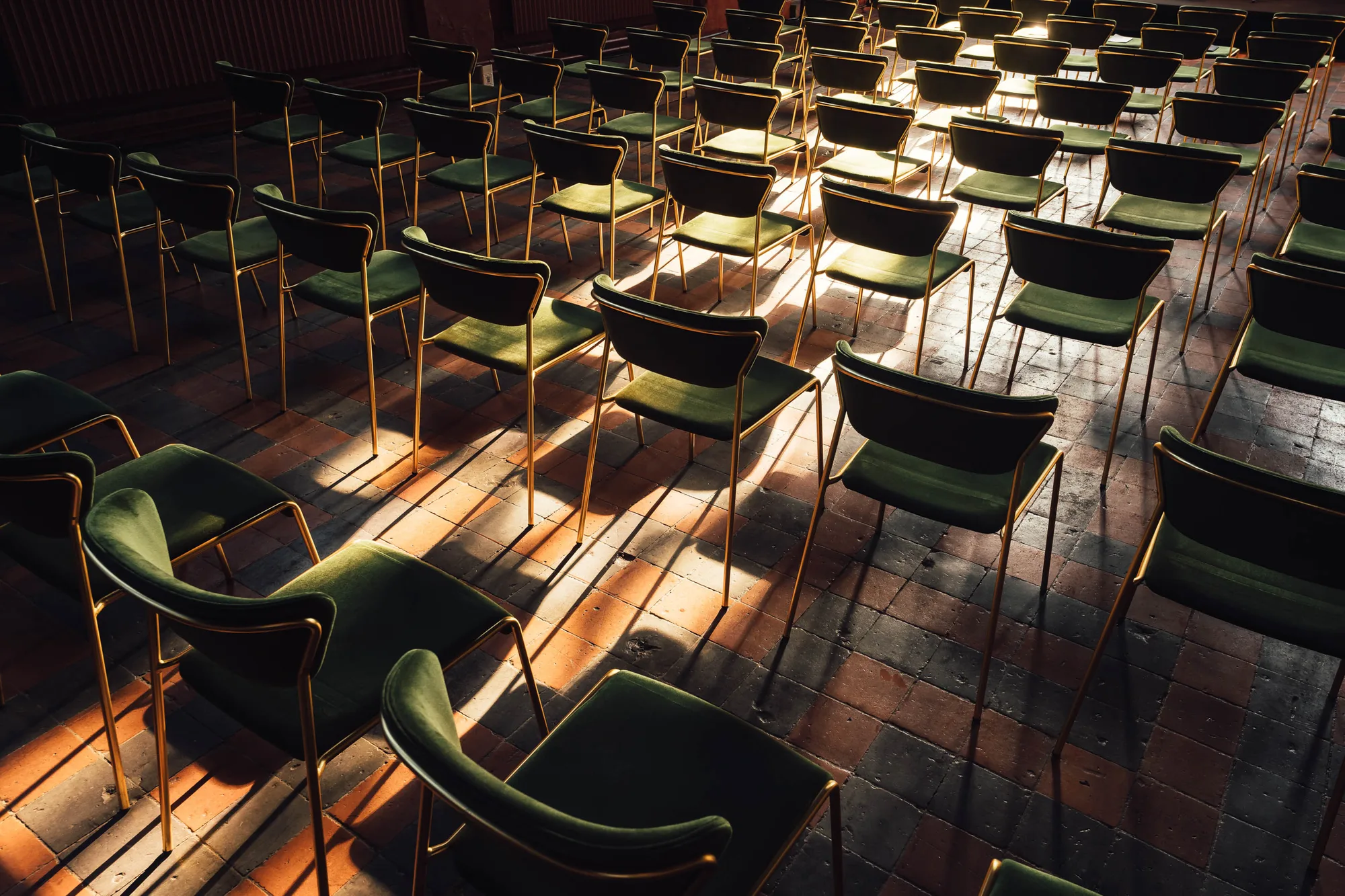 Rows of green velvet chairs with gold frames arranged on a tiled floor with sunlight casting long shadows.