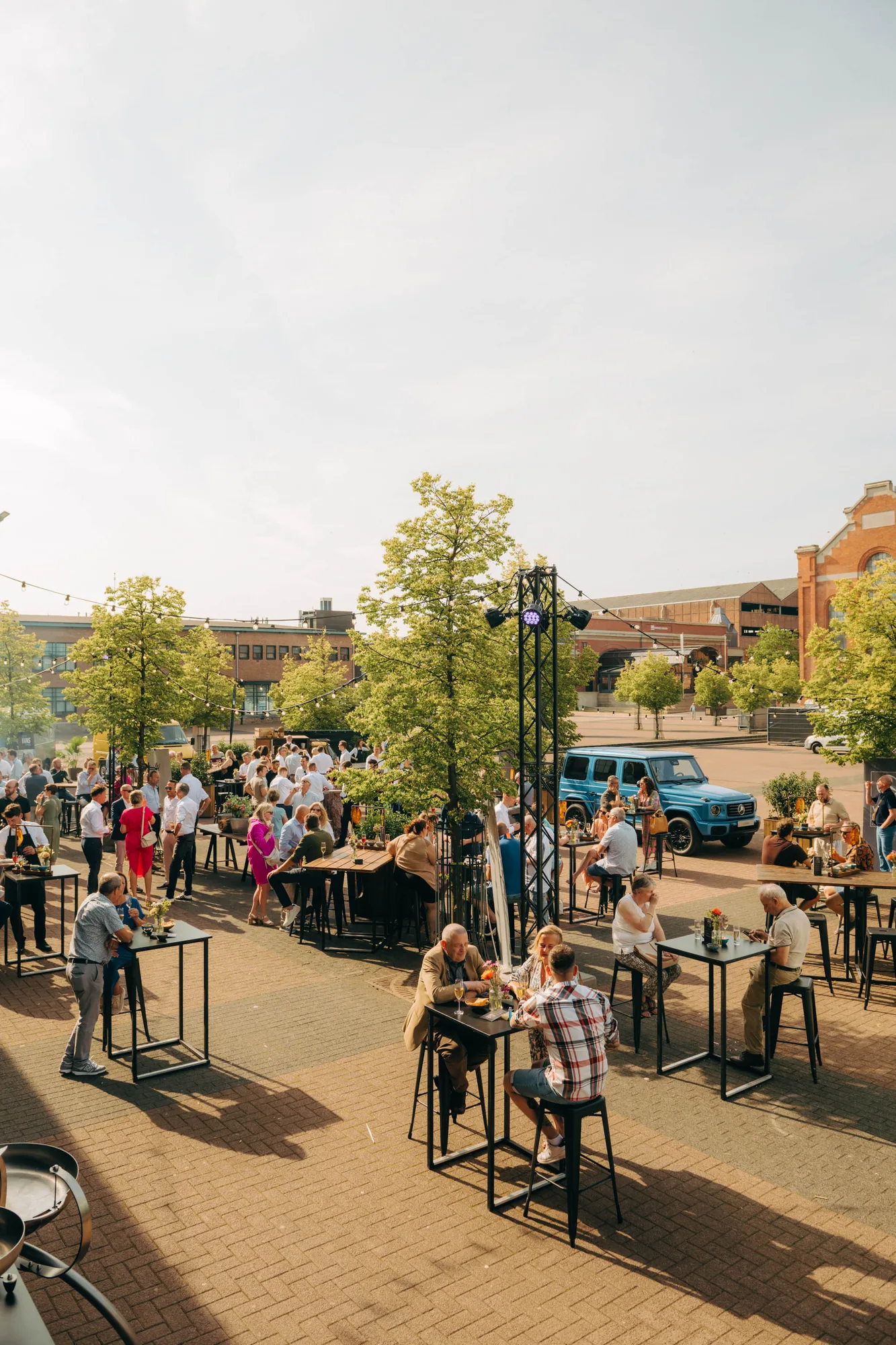 Outdoor social gathering with people sitting and standing around high tables under string lights on a sunny day.