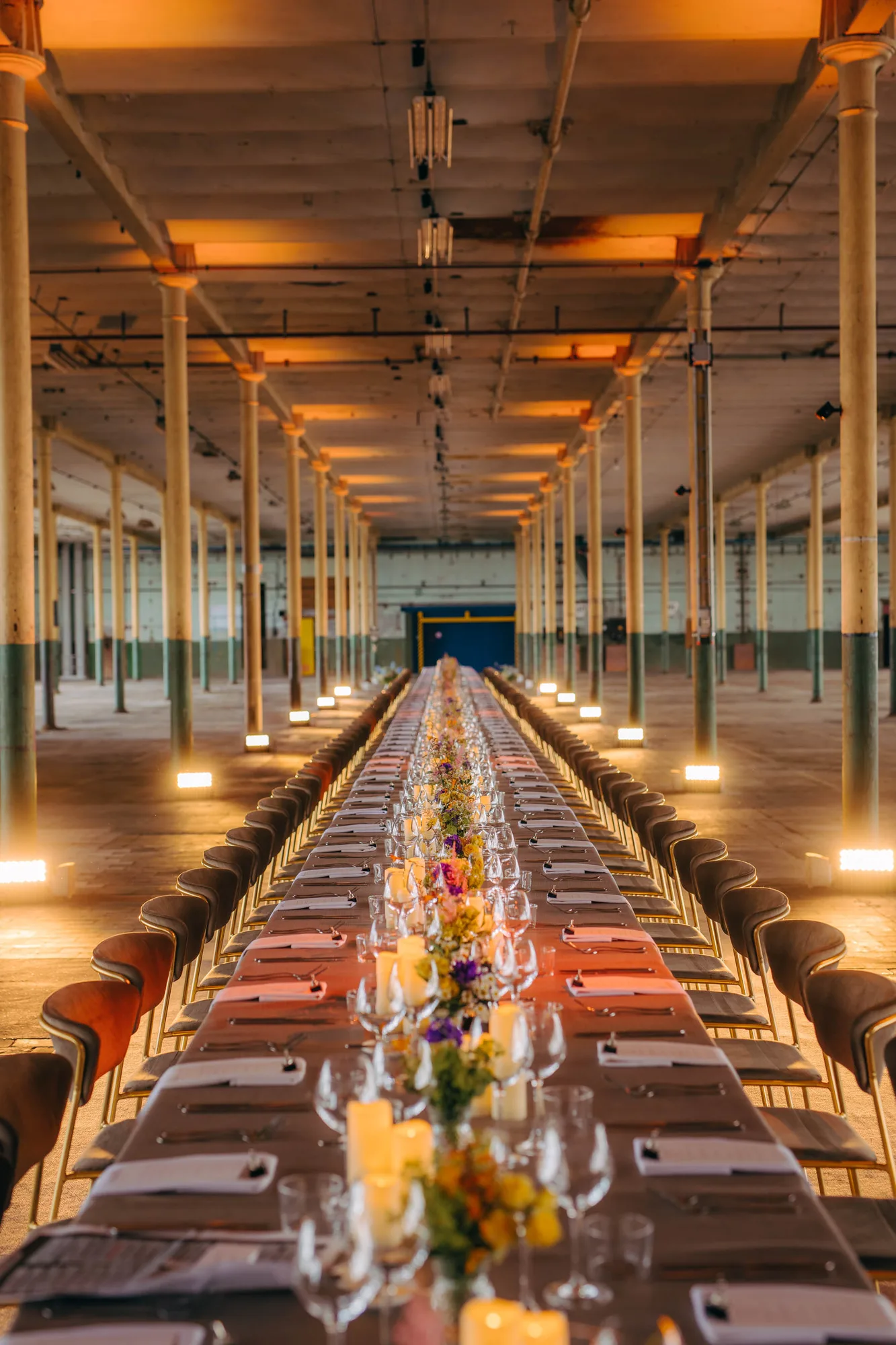 Long banquet table set with candles, flowers, wine glasses, and menus in a large industrial-style hall with columns.