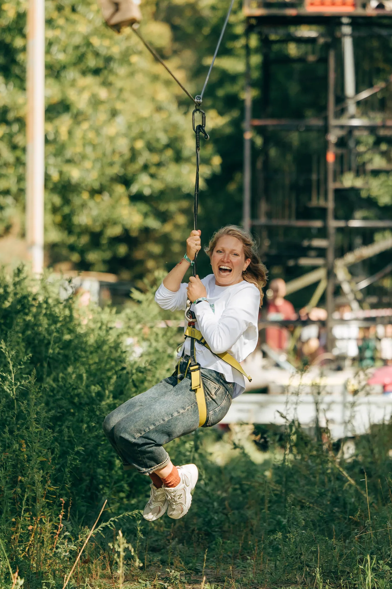 Smiling woman in a white long-sleeve shirt and jeans riding a zipline over green foliage.