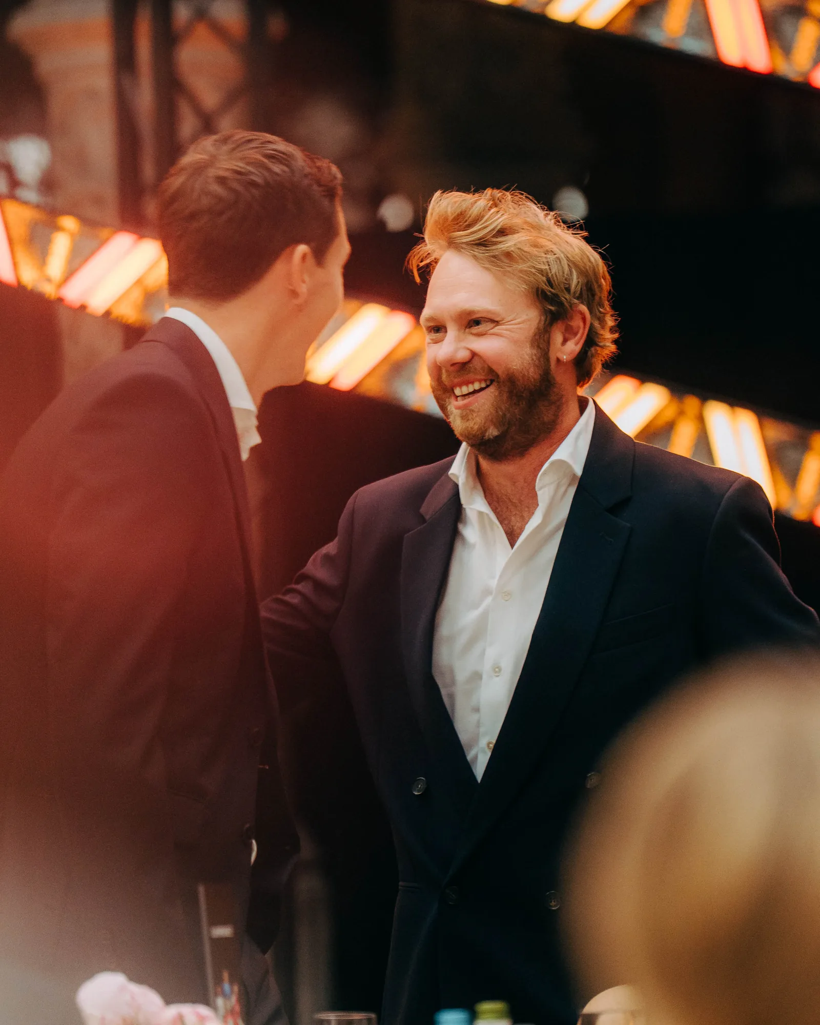 Two men in dark suits smiling and talking warmly at an indoor event with festive lighting in the background.