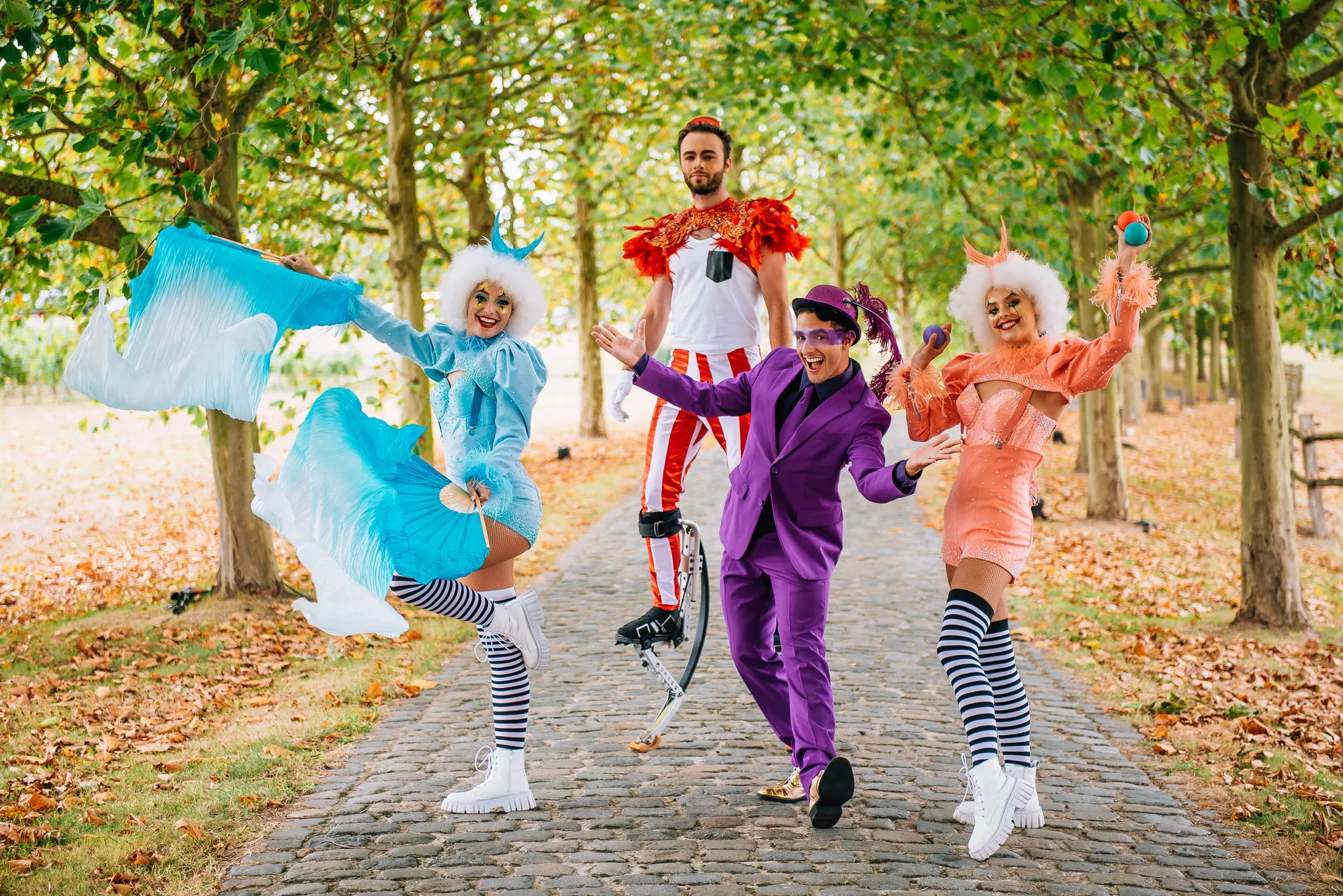 Four performers in colorful costumes on a cobblestone path lined with trees, including one on stilts and two with striped stockings.