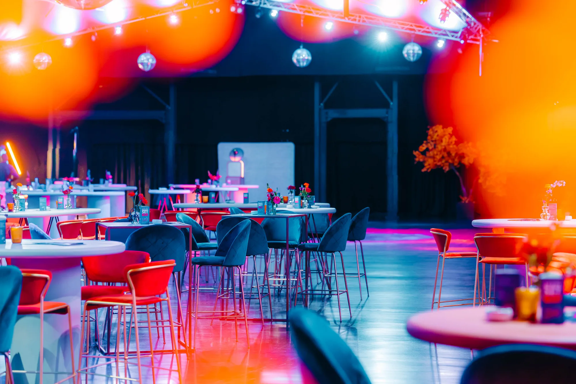 Empty modern event space with colorful lighting, featuring round tables and red and blue chairs.
