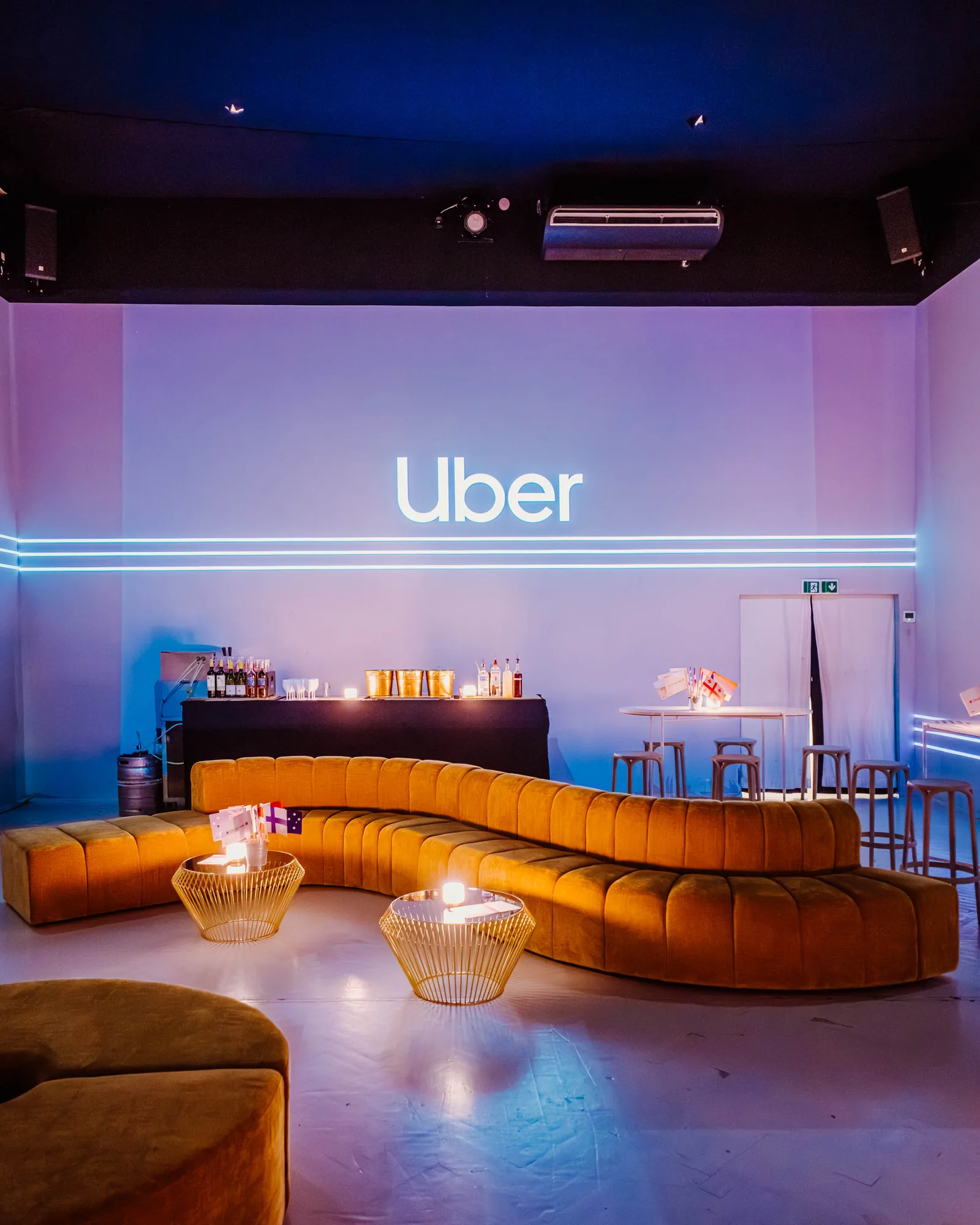 Modern lounge area with curved mustard-yellow sofa, round gold wireframe tables with candles, and illuminated Uber logo on wall.