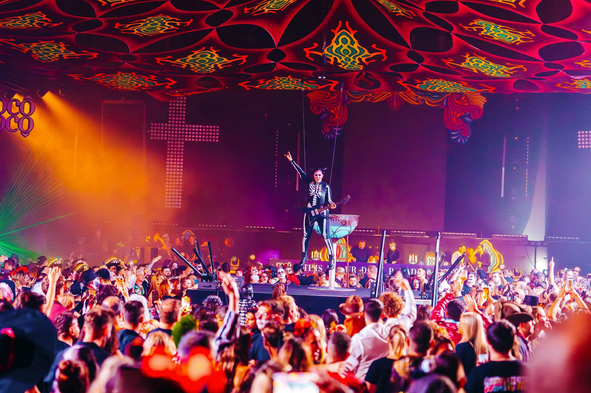 Crowded nightclub with a performer in a skeleton costume playing guitar on a raised platform under colorful patterned ceiling.