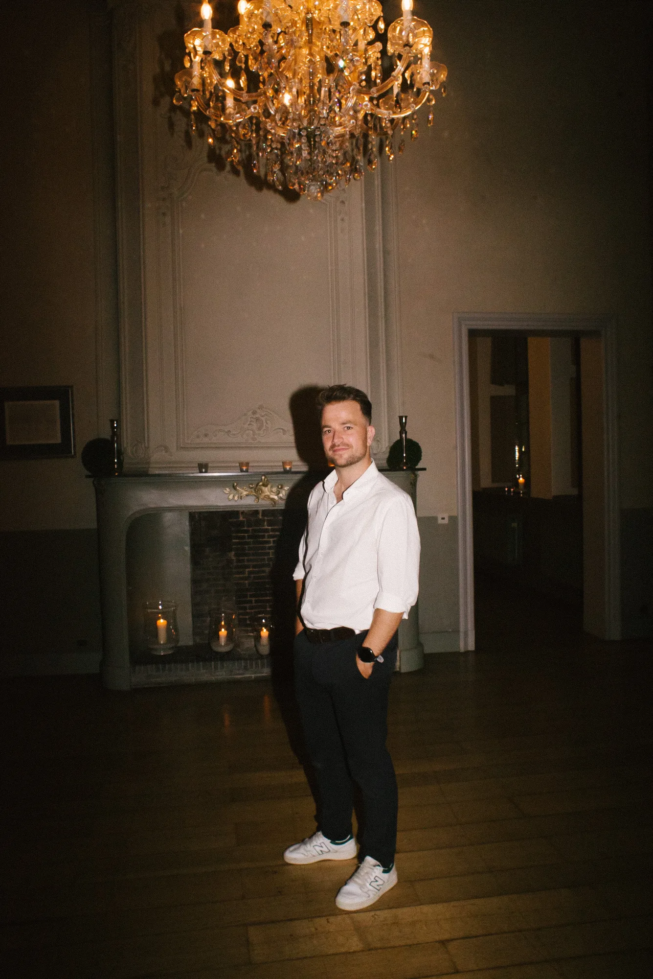 Man in white shirt and dark pants standing in a dimly lit room with a chandelier and candles in a fireplace behind him.