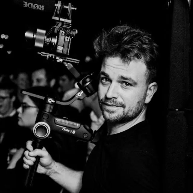 Man with short hair and beard holding a professional camera mounted on a Crane 3S gimbal stabilizer in a dark setting.