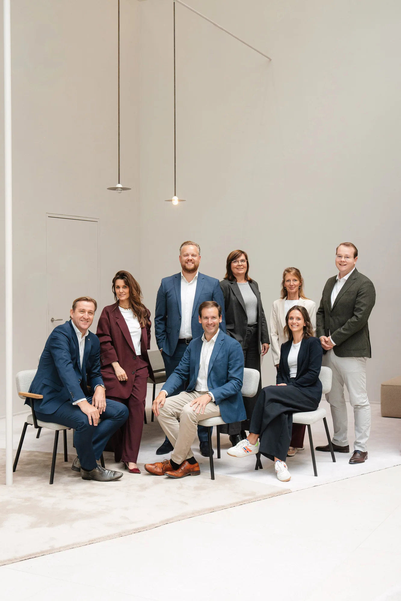 Group of eight professionally dressed people posing indoors with three seated on chairs and five standing behind them.