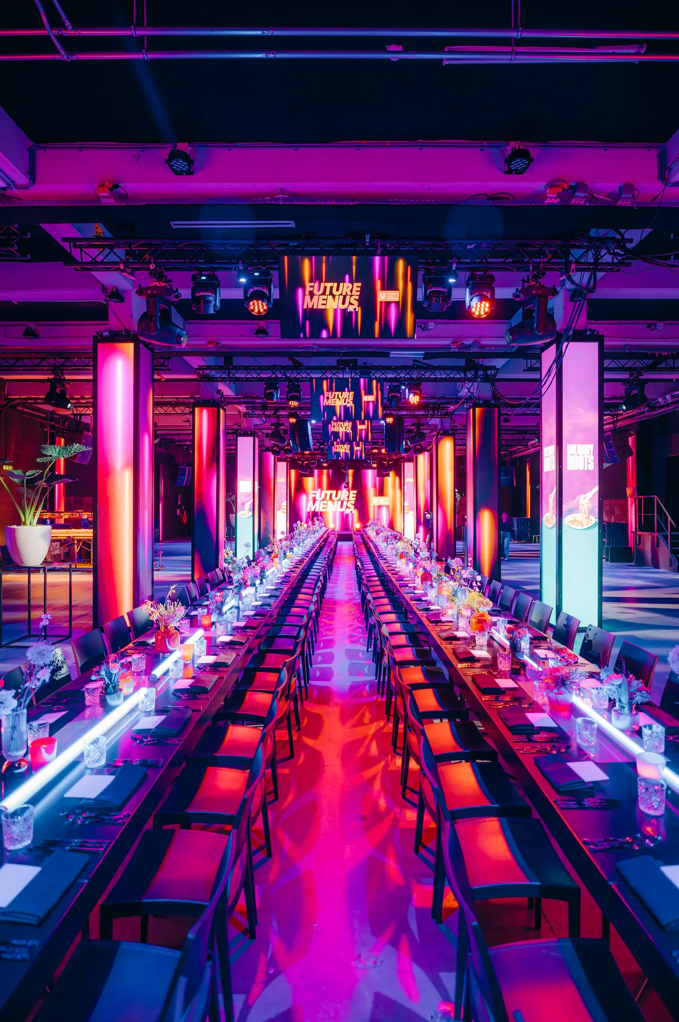 Long banquet tables with black chairs and floral centerpieces under bright pink and orange neon lights with screens displaying 'Future Menus'.
