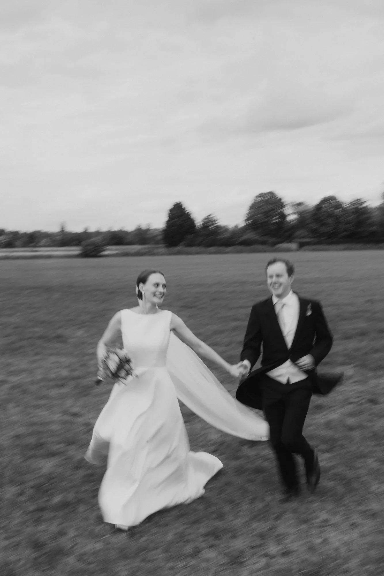 Black and white photo of a bride and groom holding hands and running across a grassy field.