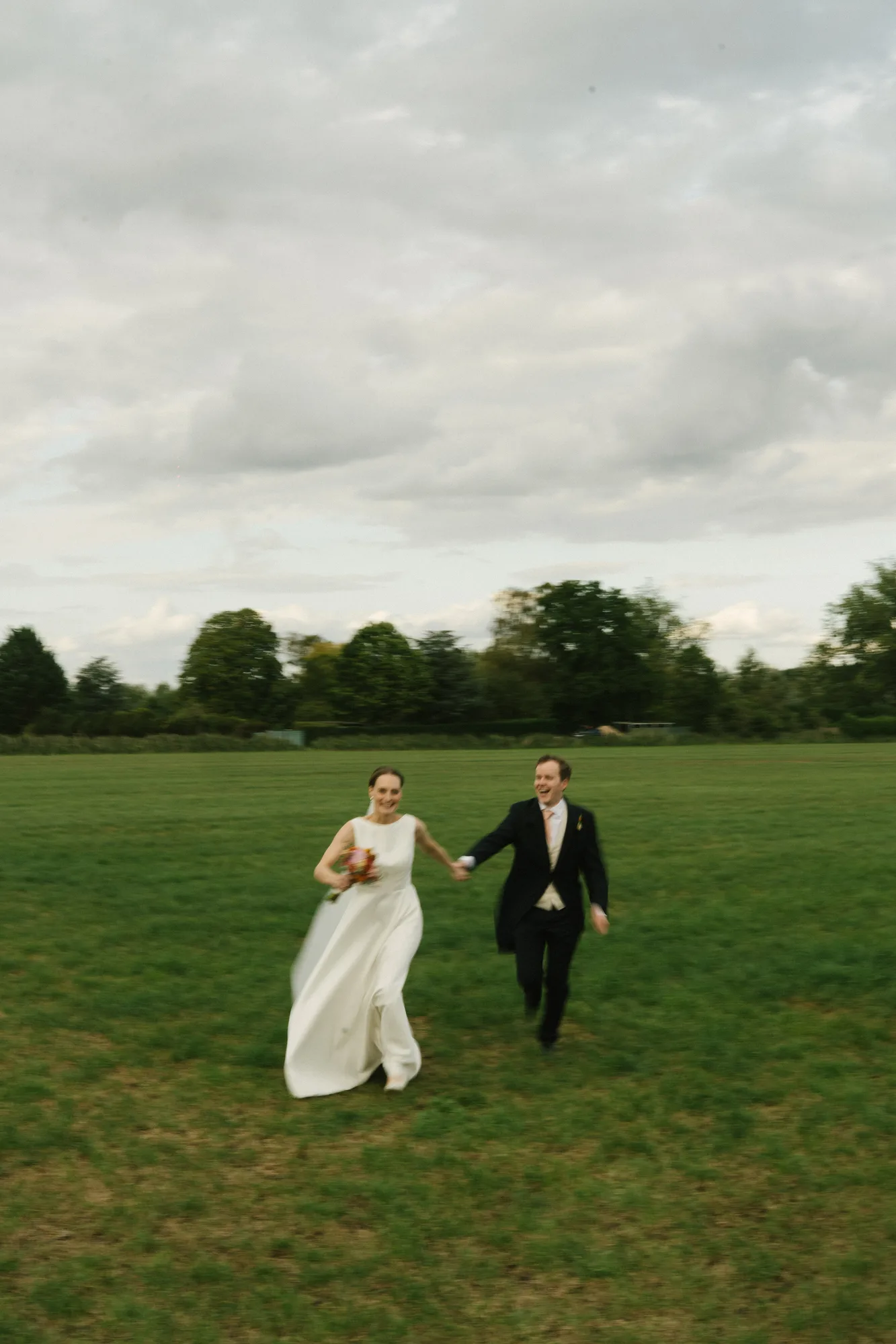 Bride and groom holding hands and running across a green field under a cloudy sky.