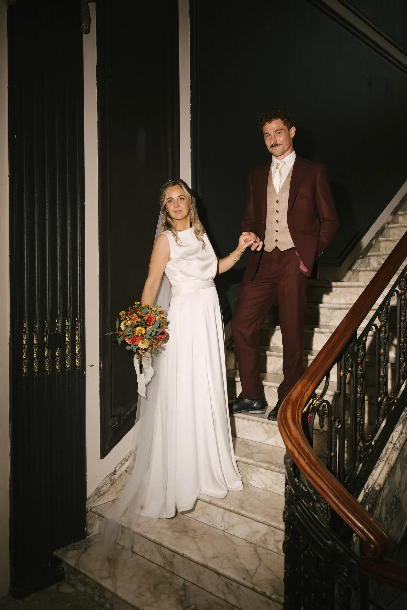 Bride in white dress holding a bouquet and groom in maroon suit standing on marble stairs holding hands.