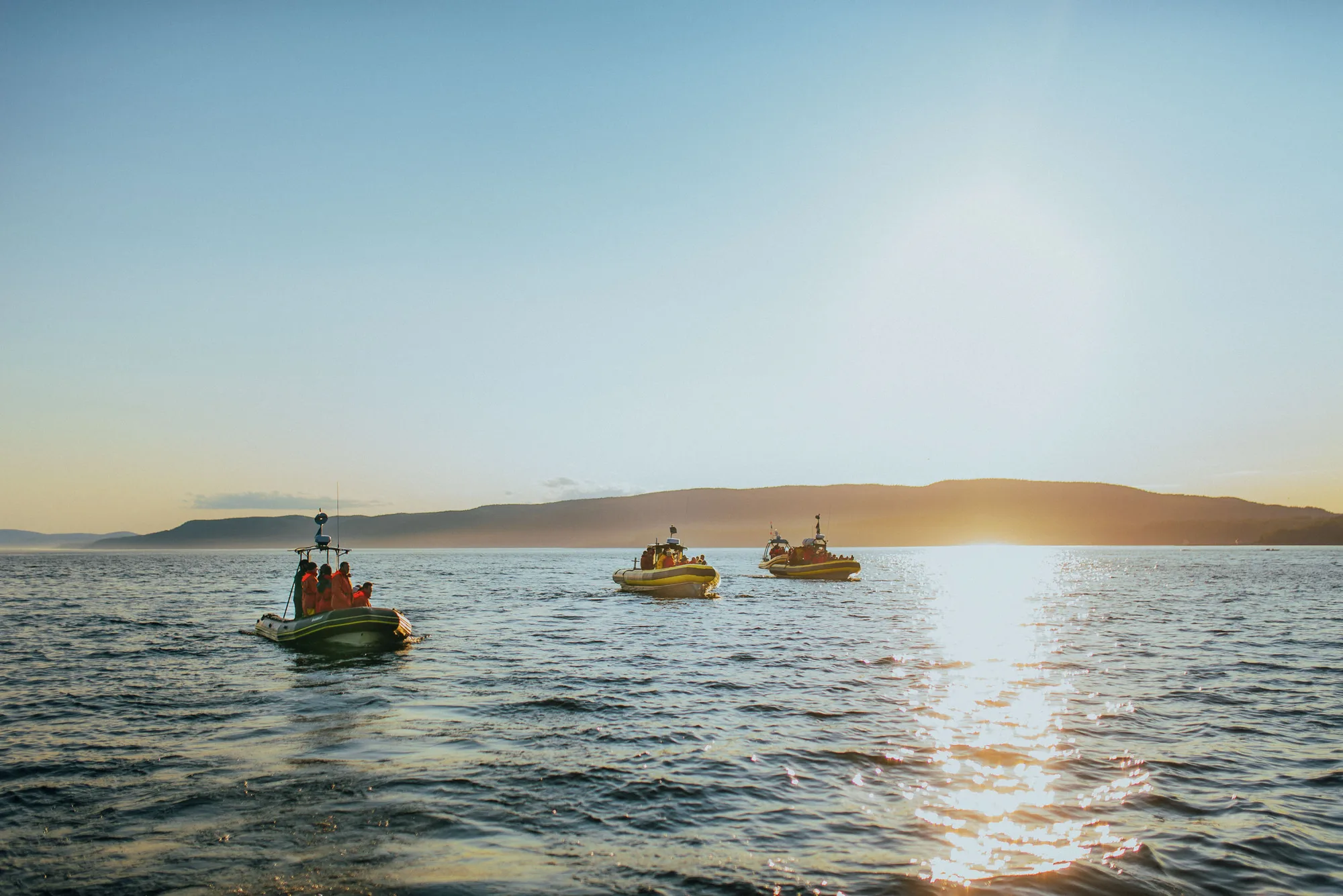 Three small boats on a calm sea at sunset with a mountainous horizon and people wearing orange jackets on board.