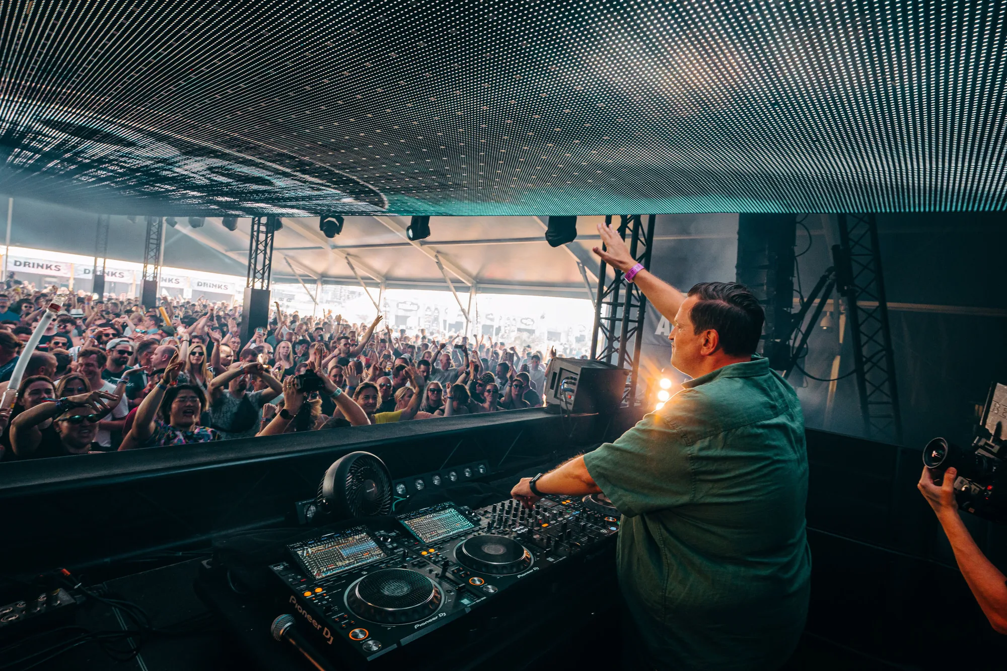 DJ performing on stage with hands raised, facing a large crowd under a lit ceiling panel at a music event.