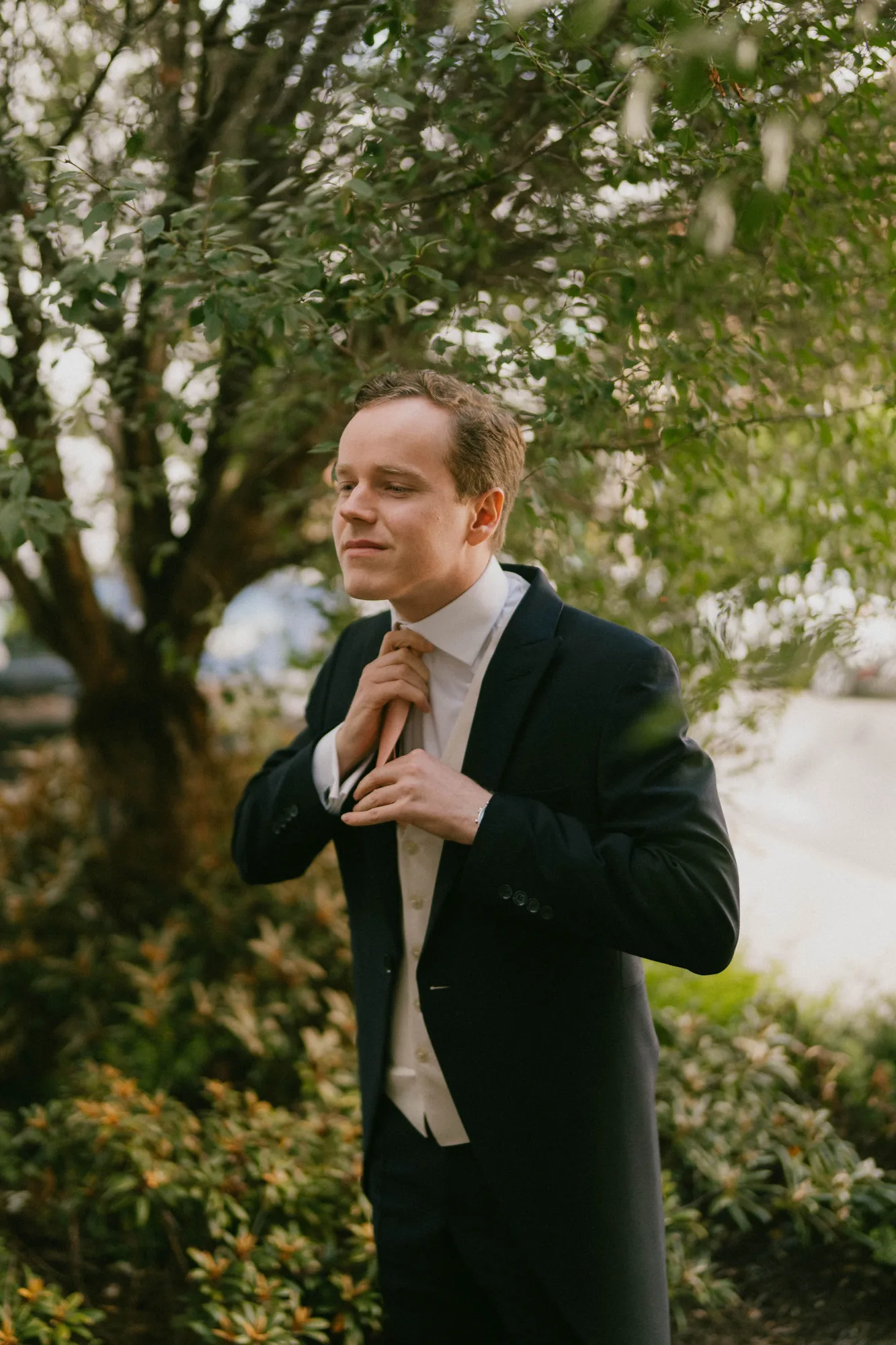 Man in a black suit and white vest adjusting his tie outdoors near green bushes and a tree.