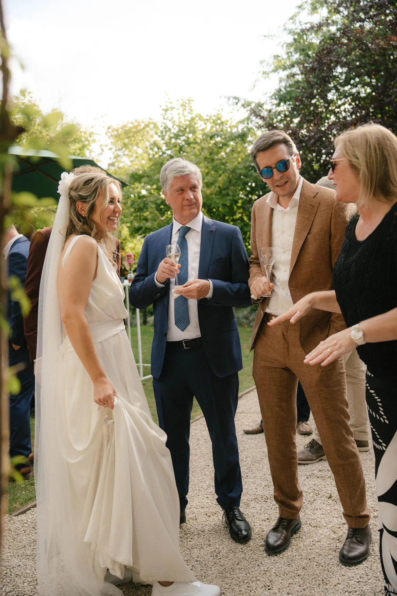 Bride in white dress and veil smiling and talking with three wedding guests outdoors, two men with glasses holding champagne flutes and a woman gesturing with her hands.