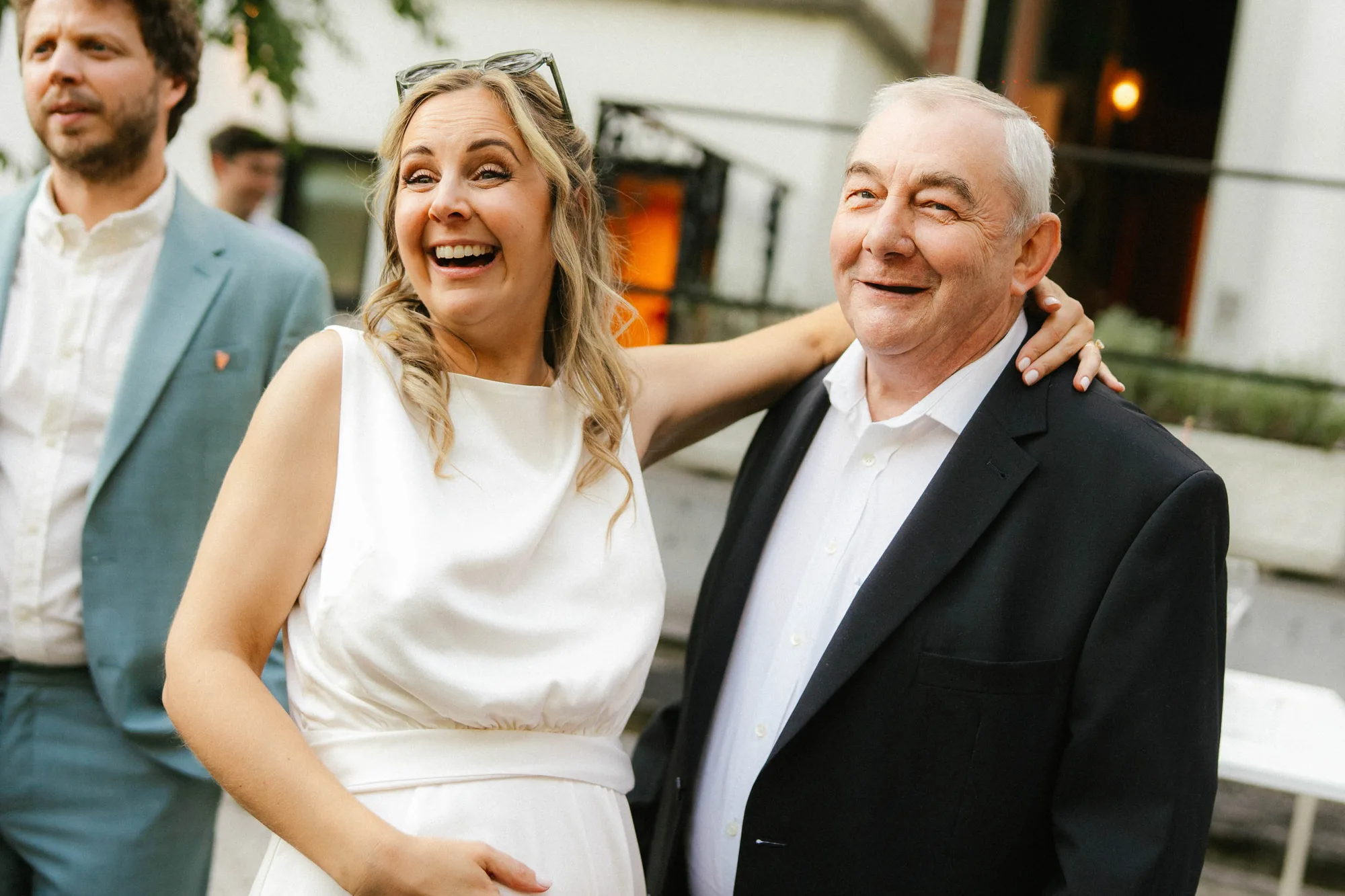 Smiling pregnant woman in white dress with arm around elderly man in a black blazer, both looking happy outdoors.