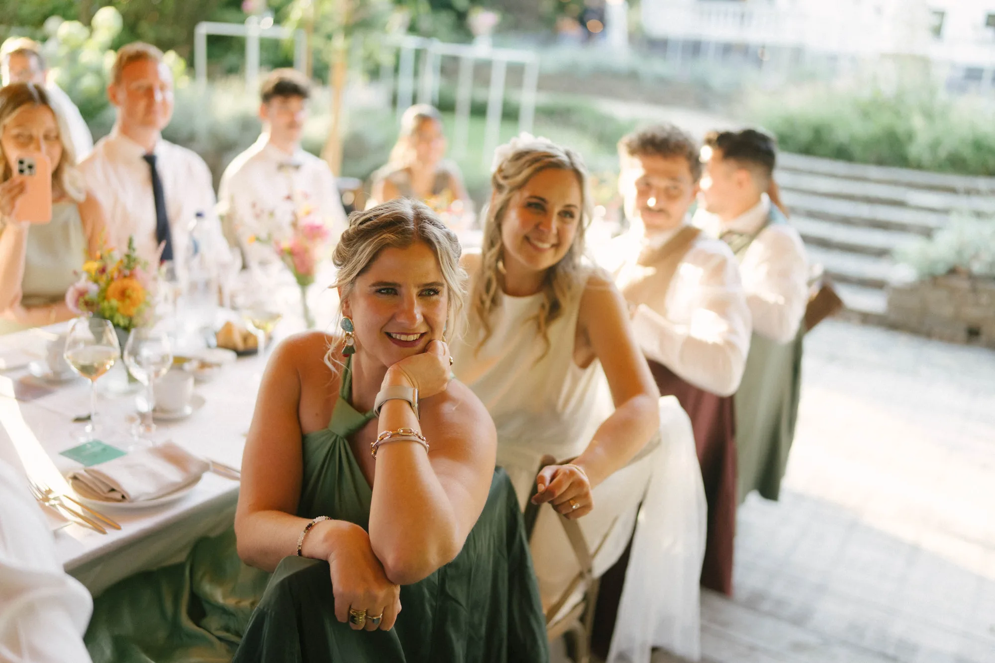 Smiling woman in green dress sitting at a table during a daytime outdoor event with guests in the background.