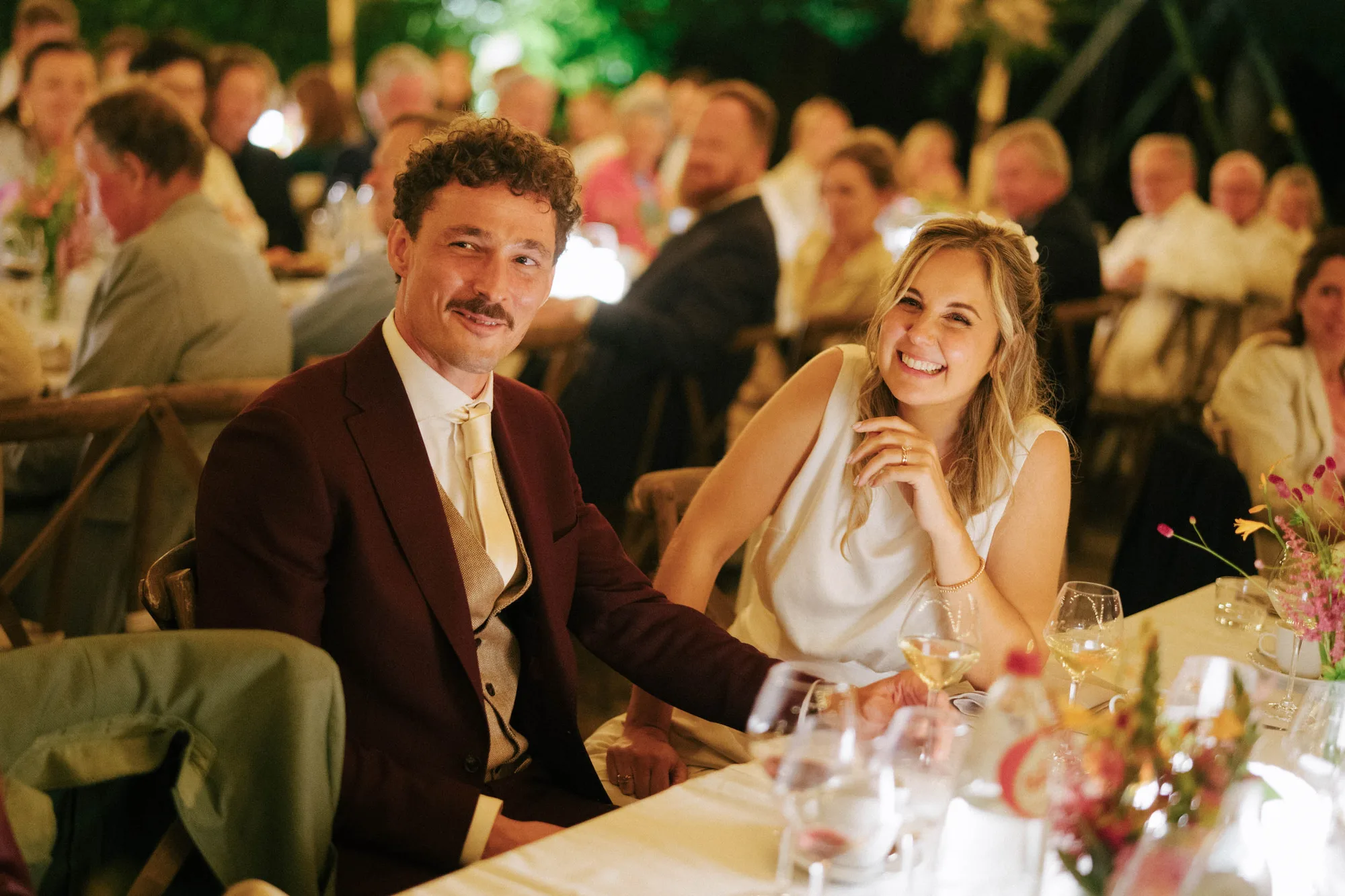 Smiling couple seated at a decorated table during an evening event with guests in the background.