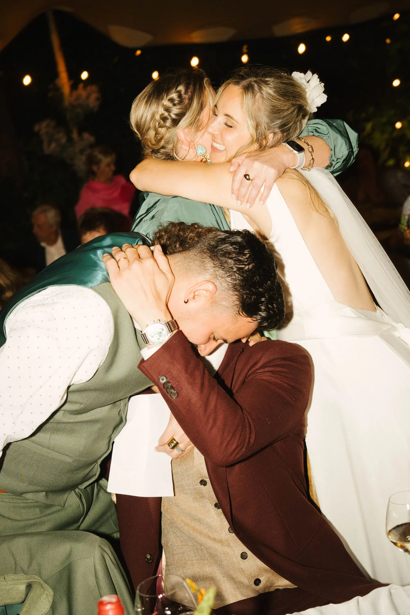 Two women warmly hugging, one in a white dress with a veil, and two men embracing closely in a joyful group hug at an event with string lights in the background.