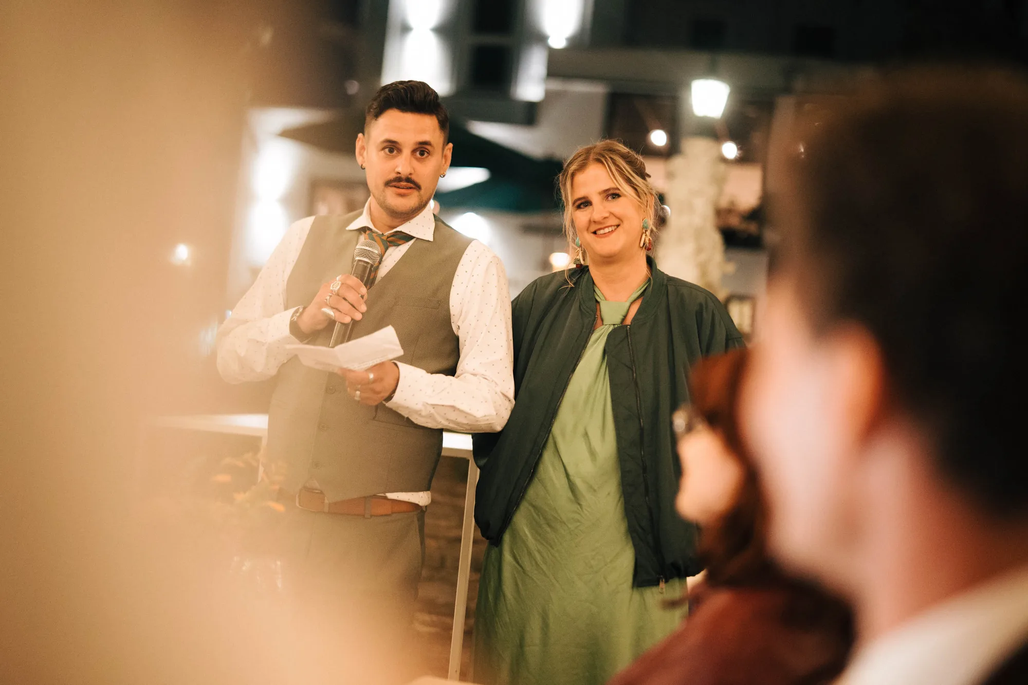 Man holding a microphone and speaking next to a smiling woman in a green dress and jacket at an indoor event.