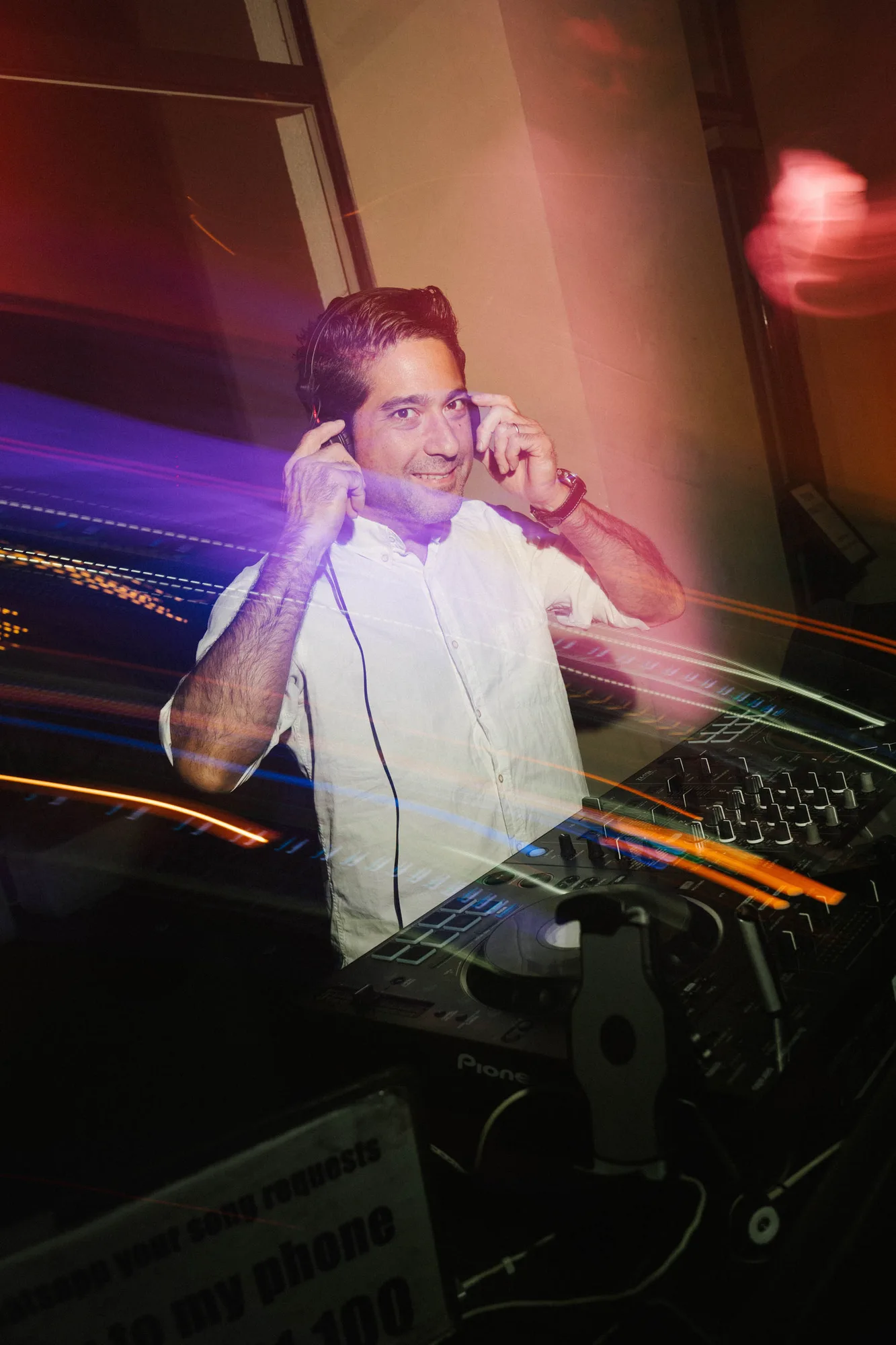 Smiling DJ wearing a white shirt adjusting headphones behind a DJ controller with colorful light trails.