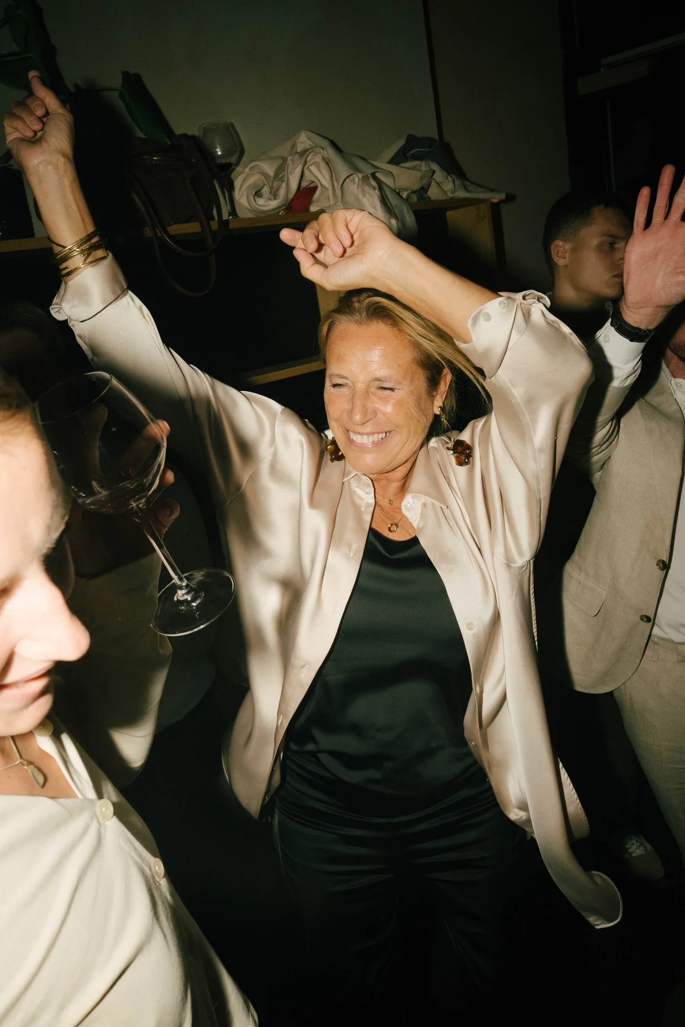 Woman in a light satin blouse smiling and dancing with arms raised at a social gathering.