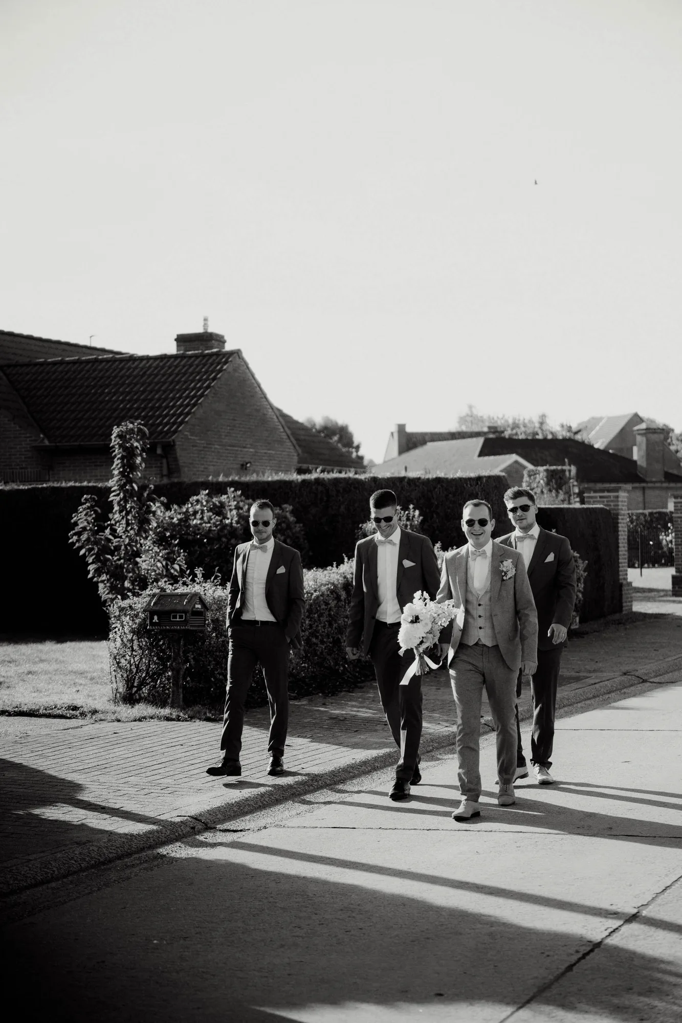 Four men dressed in suits and sunglasses walking outside on a sunny day, one holding a bouquet of flowers.
