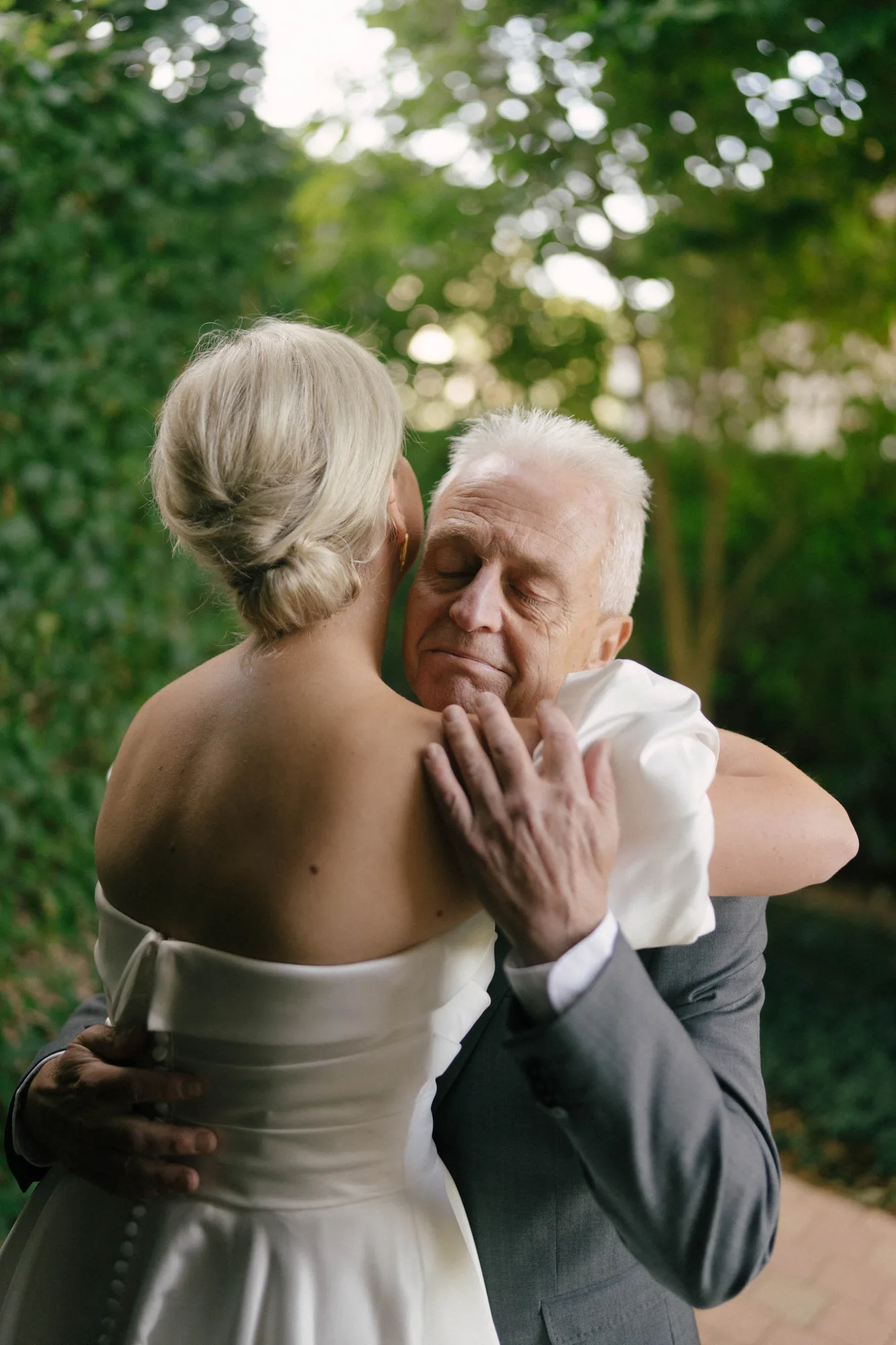 Older man in a suit hugging a woman in a white strapless dress outdoors with greenery in the background.