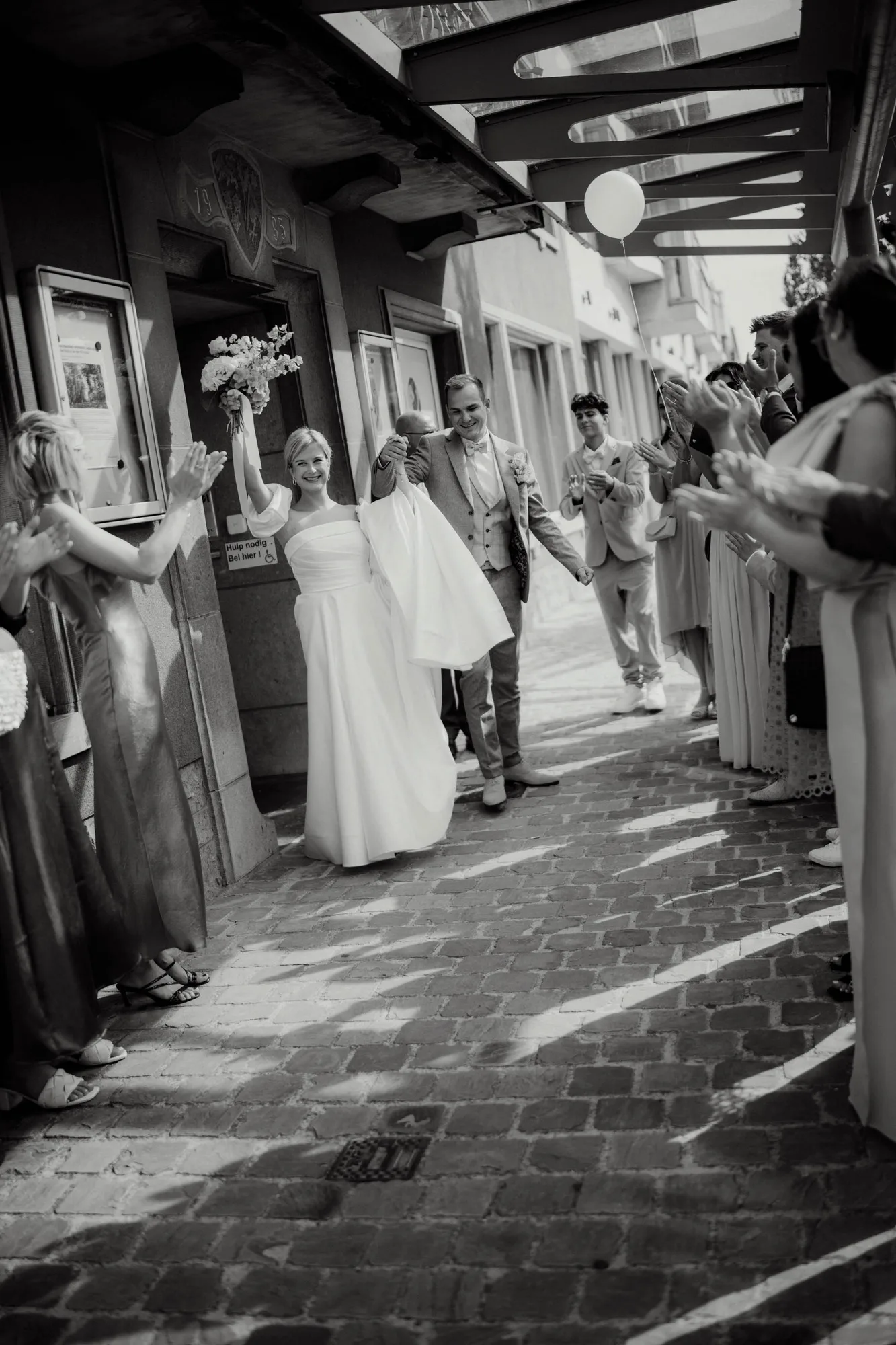 Bride and groom smiling and holding hands while walking on a cobblestone street surrounded by applauding guests.