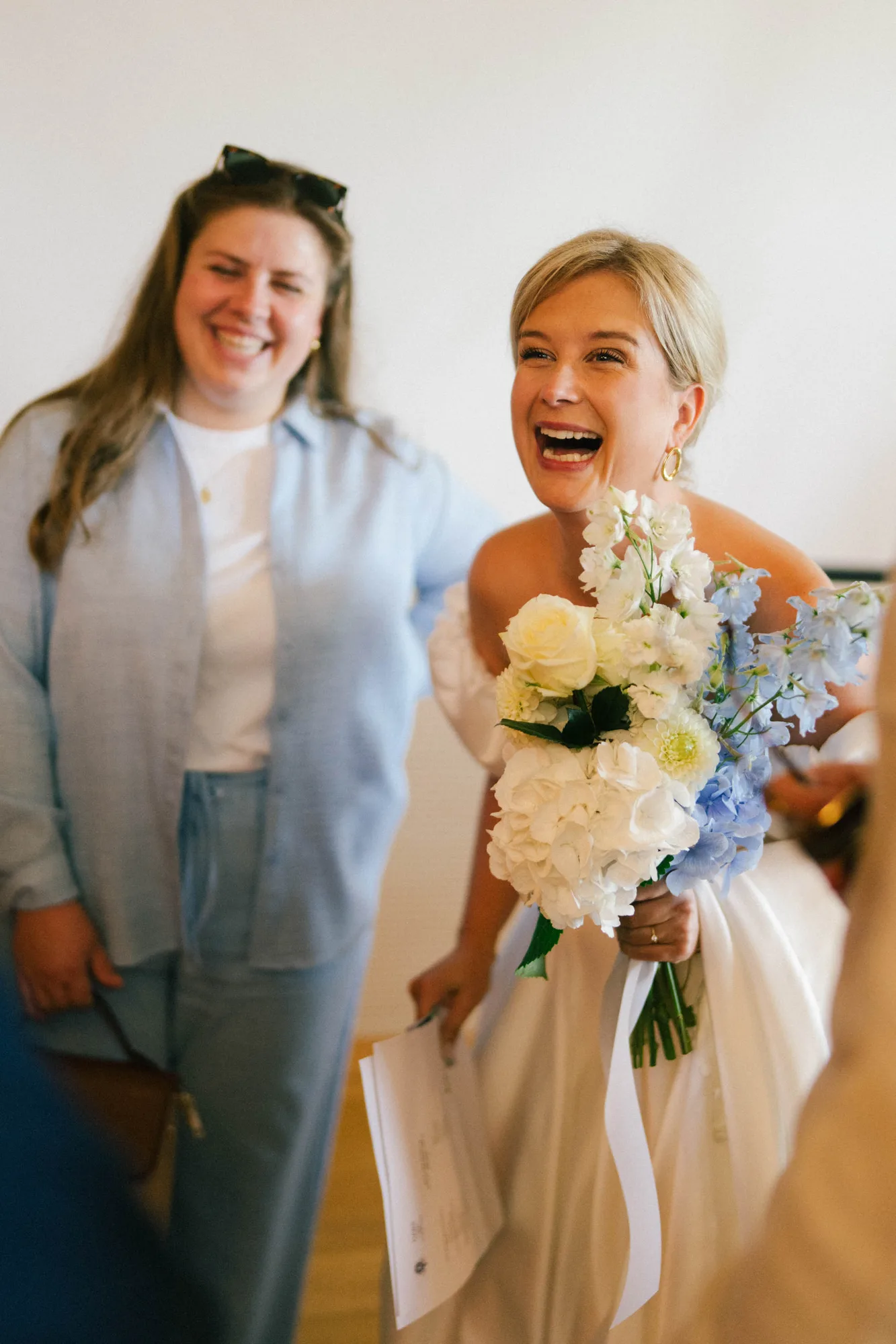 Smiling bride in white dress holding a bouquet of white and blue flowers and a paper, with a woman in casual blue shirt and jeans laughing beside her.