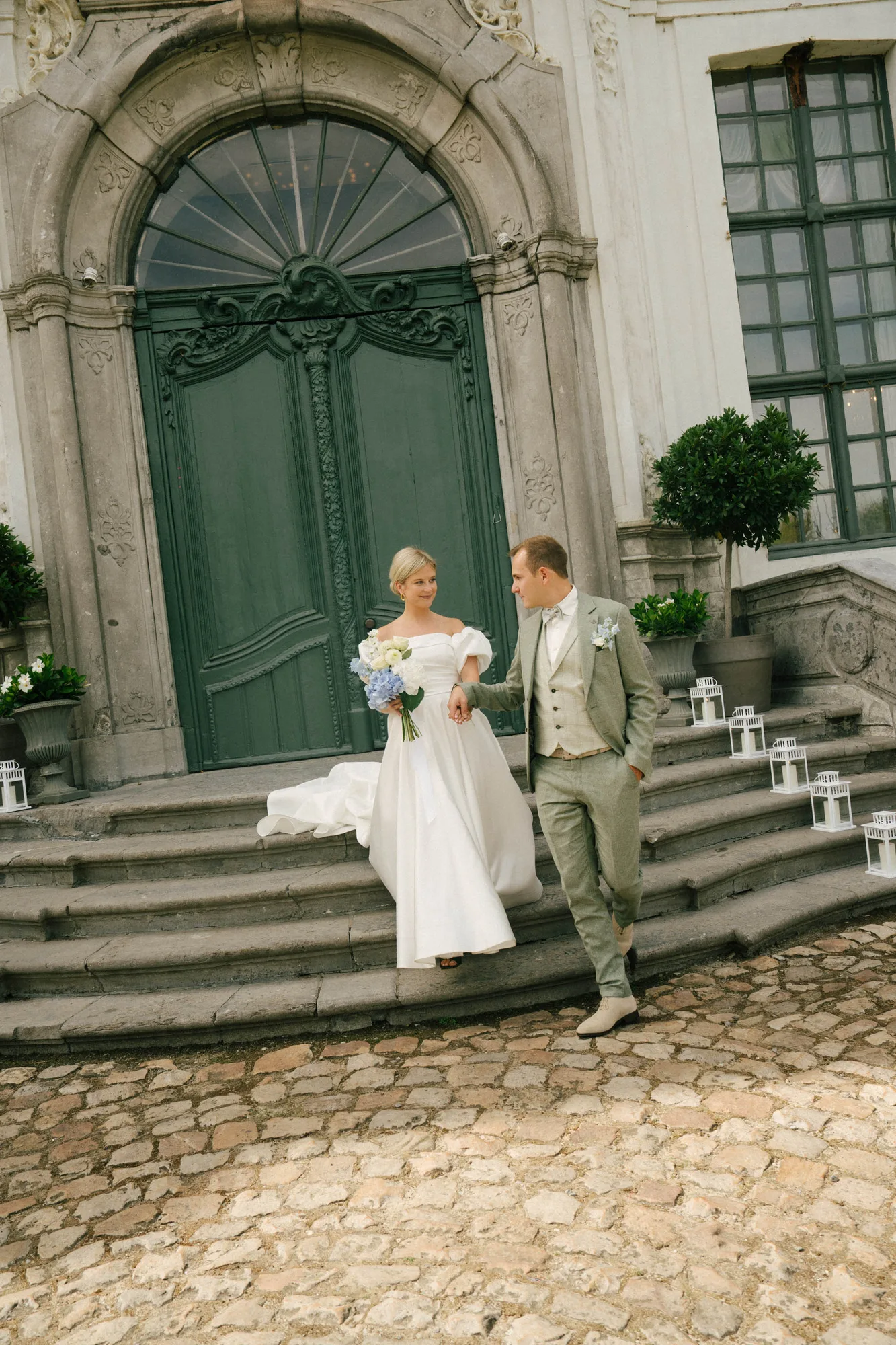 Bride in white dress holding flowers and groom in light gray suit walking down stone steps outside ornate green door.