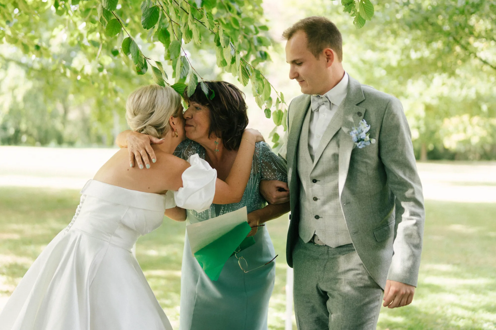 Bride in a white gown hugging and kissing an older woman in a teal dress, with a man in a gray suit standing beside them outdoors.