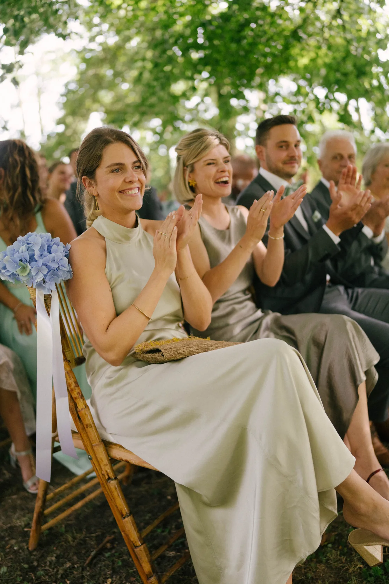 Guests sitting outdoors on wooden chairs under trees, smiling and clapping at an event.