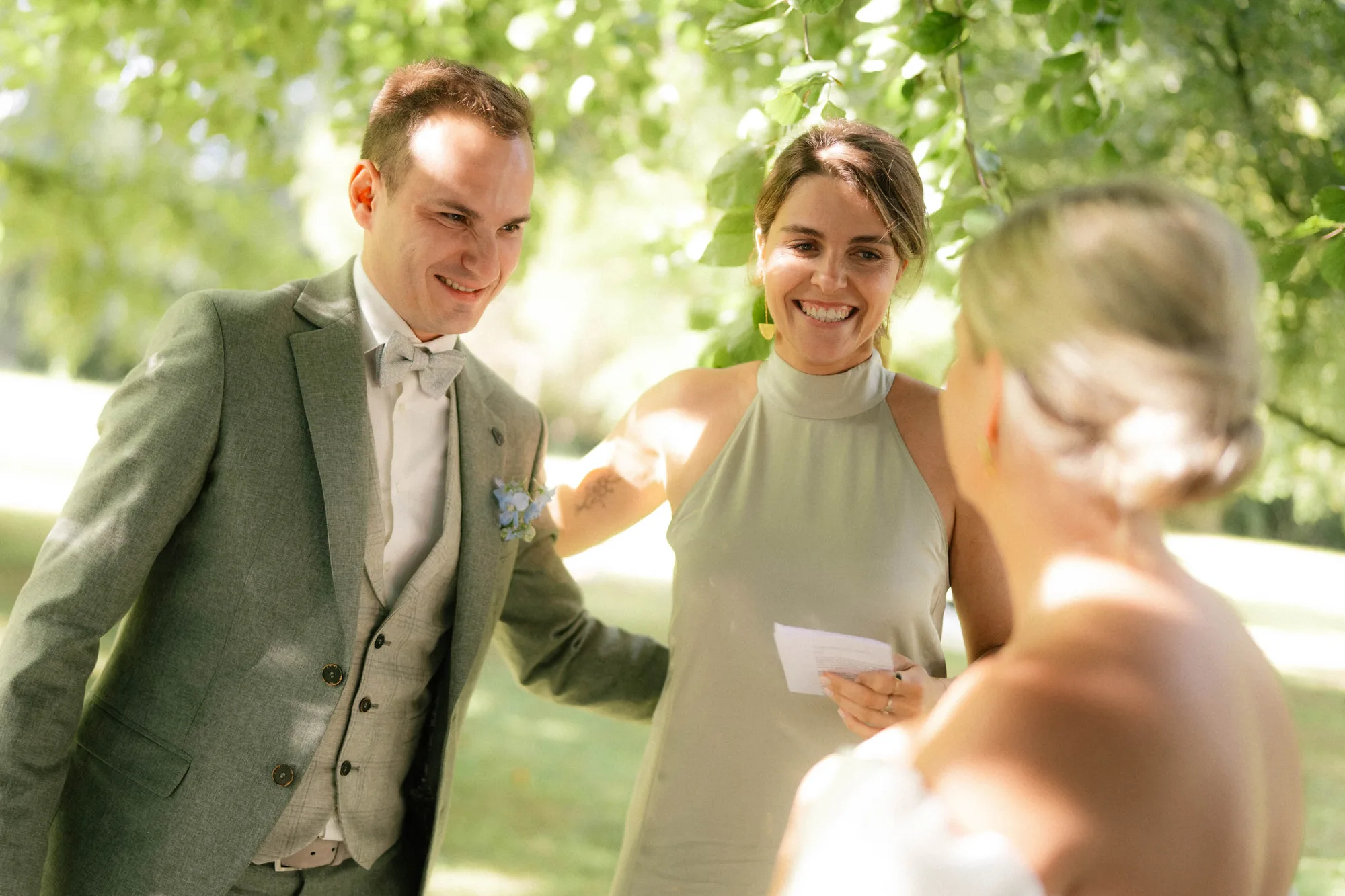 Smiling woman in a sleeveless dress holding a note stands with a man in a suit and bow tie outdoors, speaking to a woman in a bridal gown.