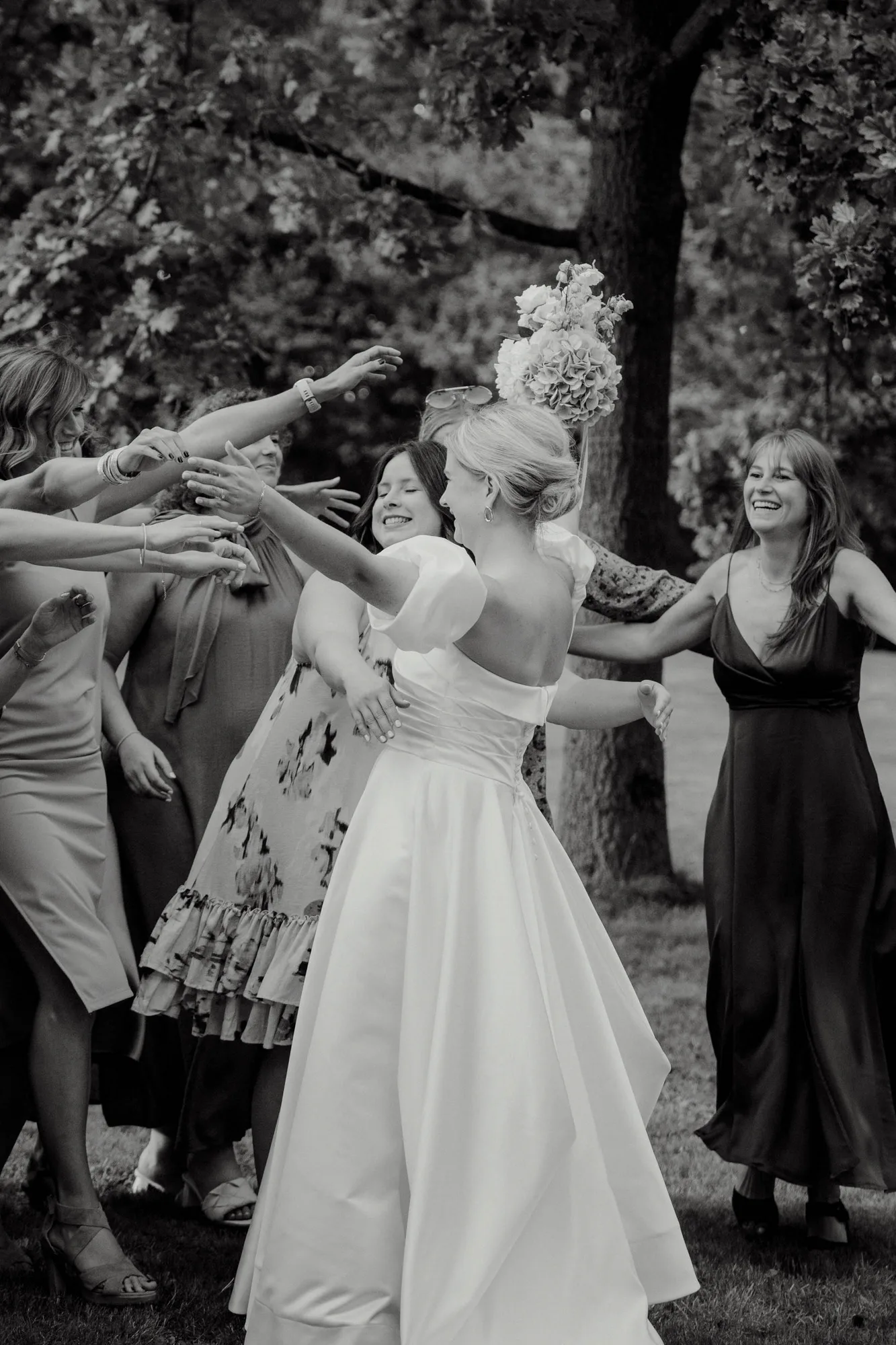 Bride in a white dress with a bouquet joyfully reaching out to a group of women at an outdoor wedding.