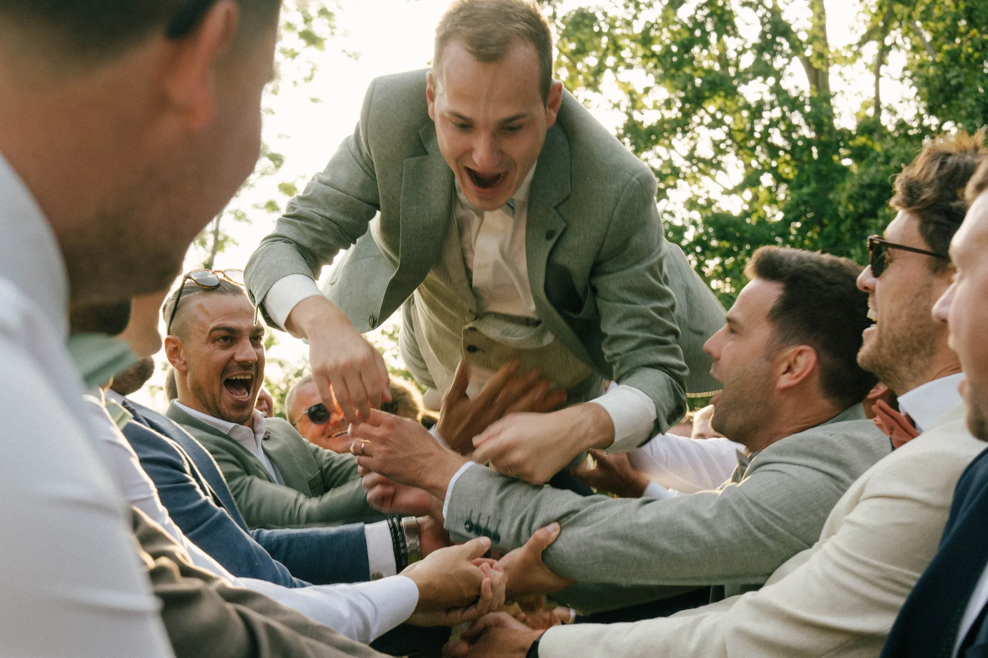 Group of men in suits lifting and cheering a man in a gray suit who is smiling and leaning forward outdoors.