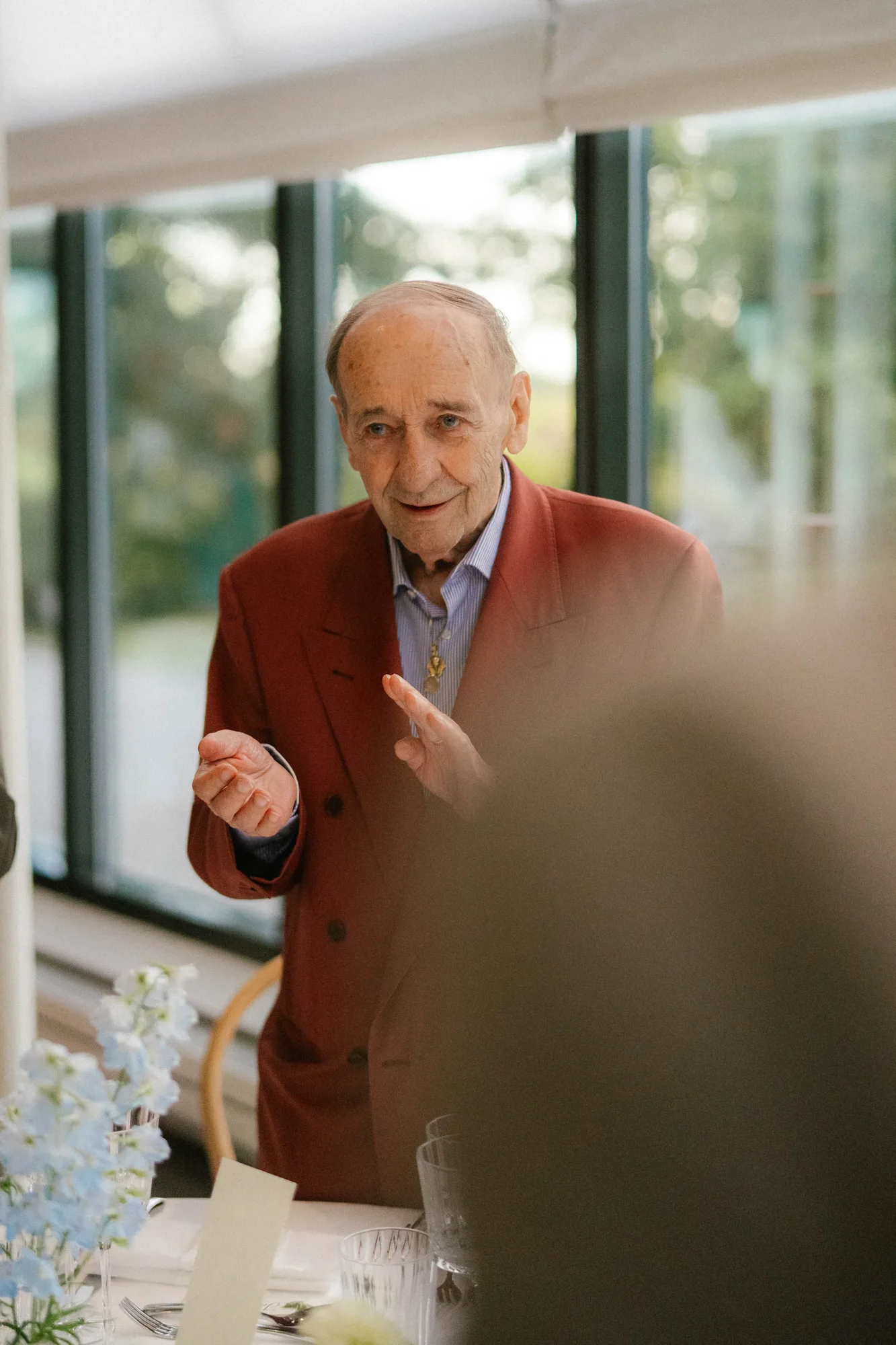 Elderly man in a rust-colored blazer speaking and gesturing at a dining table near large windows.