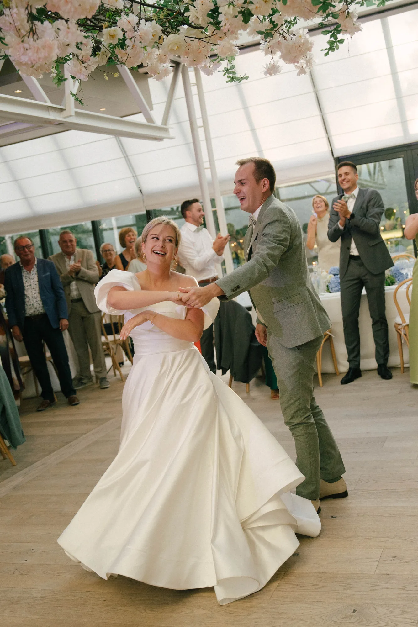 Bride and groom smiling and dancing happily indoors with guests clapping in the background.